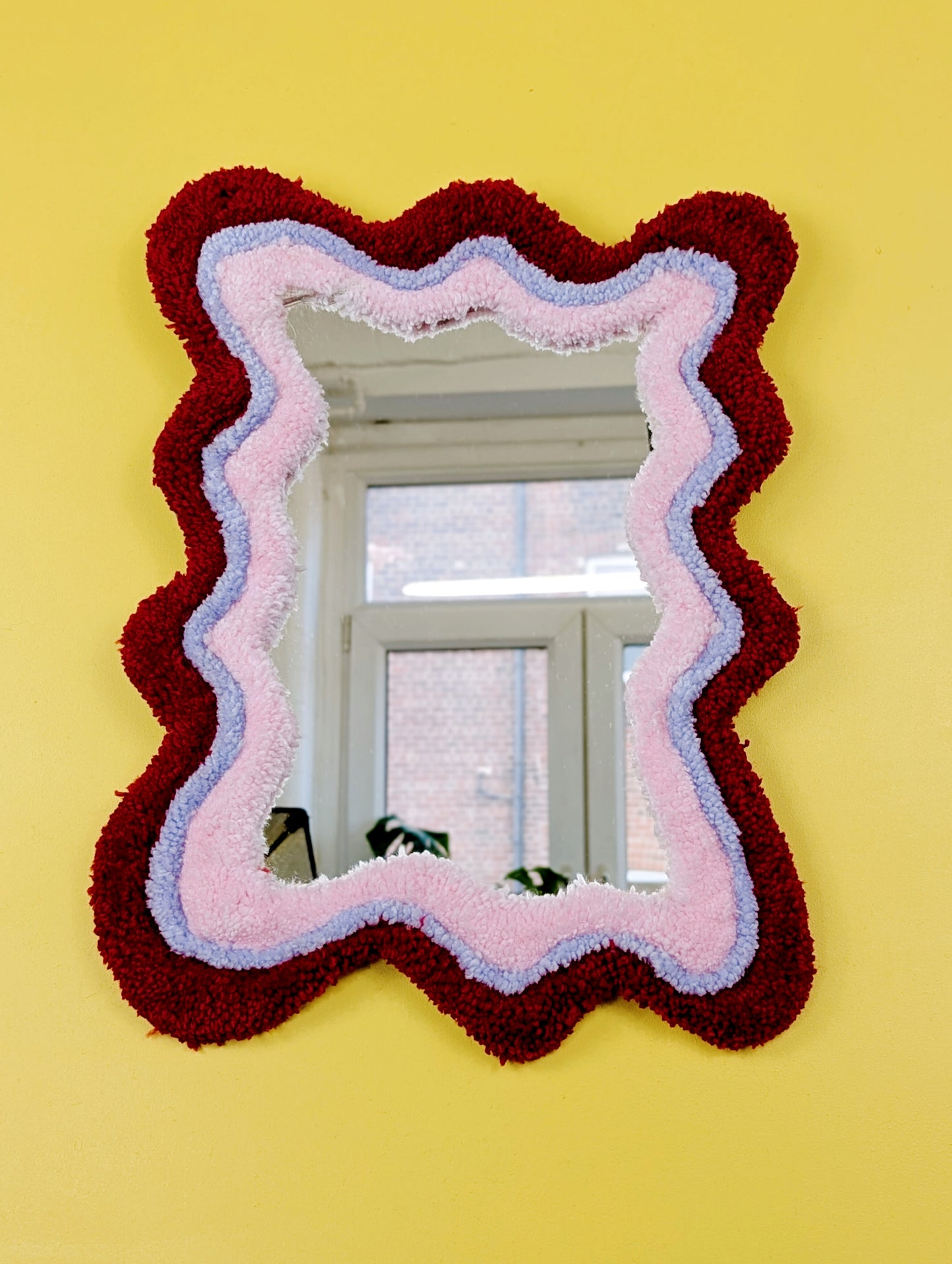Handmade wavy tufted mirror with colourful textured frame in alternating shades of bright red, pale purple and pale pink, modern aesthetic wall décor for home interiors.