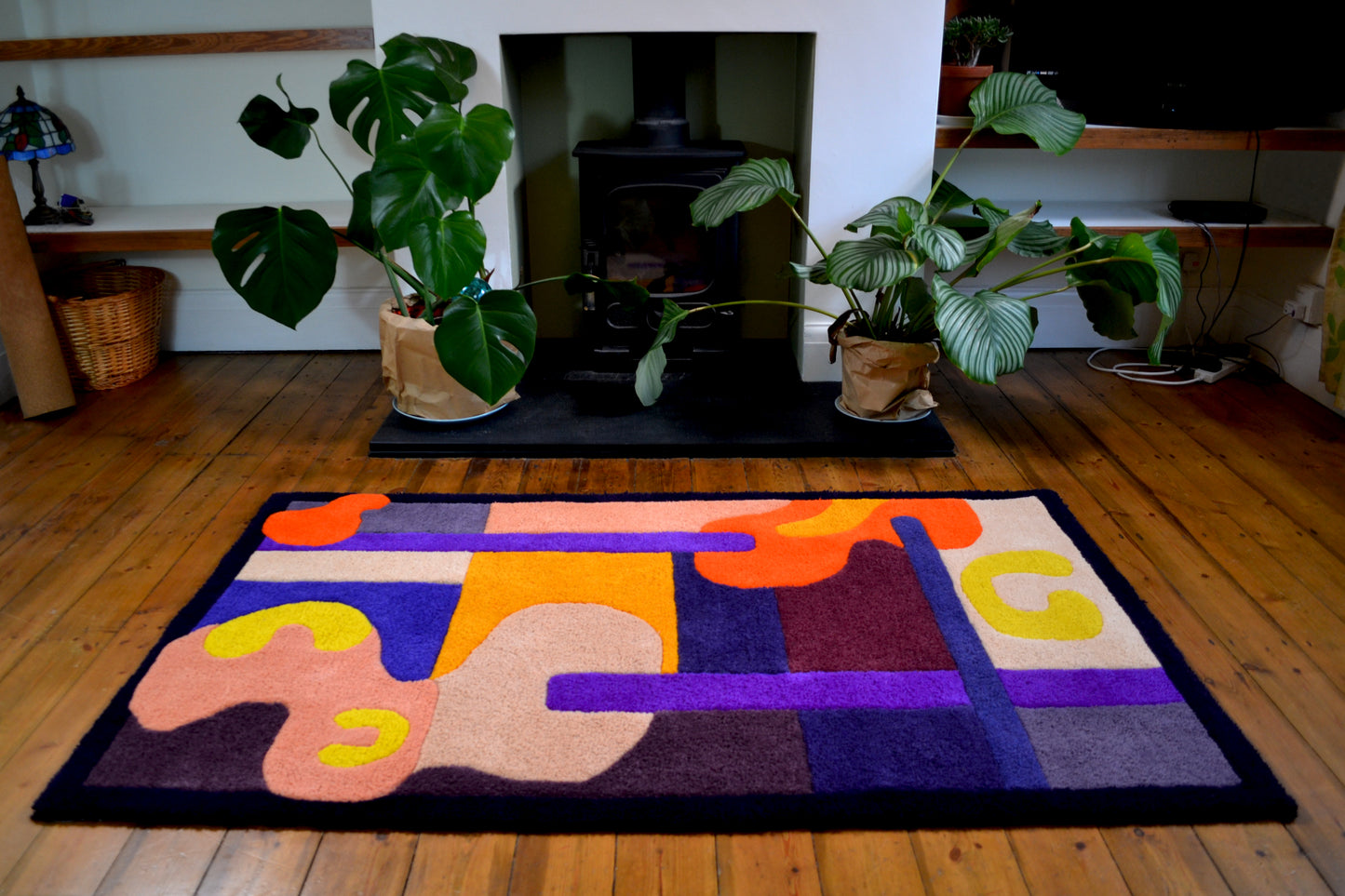 Image shows a large purple, orange, yellow, cream and burgundy handcrafted tufted wool rug, positioned in front of a log burner fireplace, with plants positioned at the sides. Rug is placed on wooden floorboards.