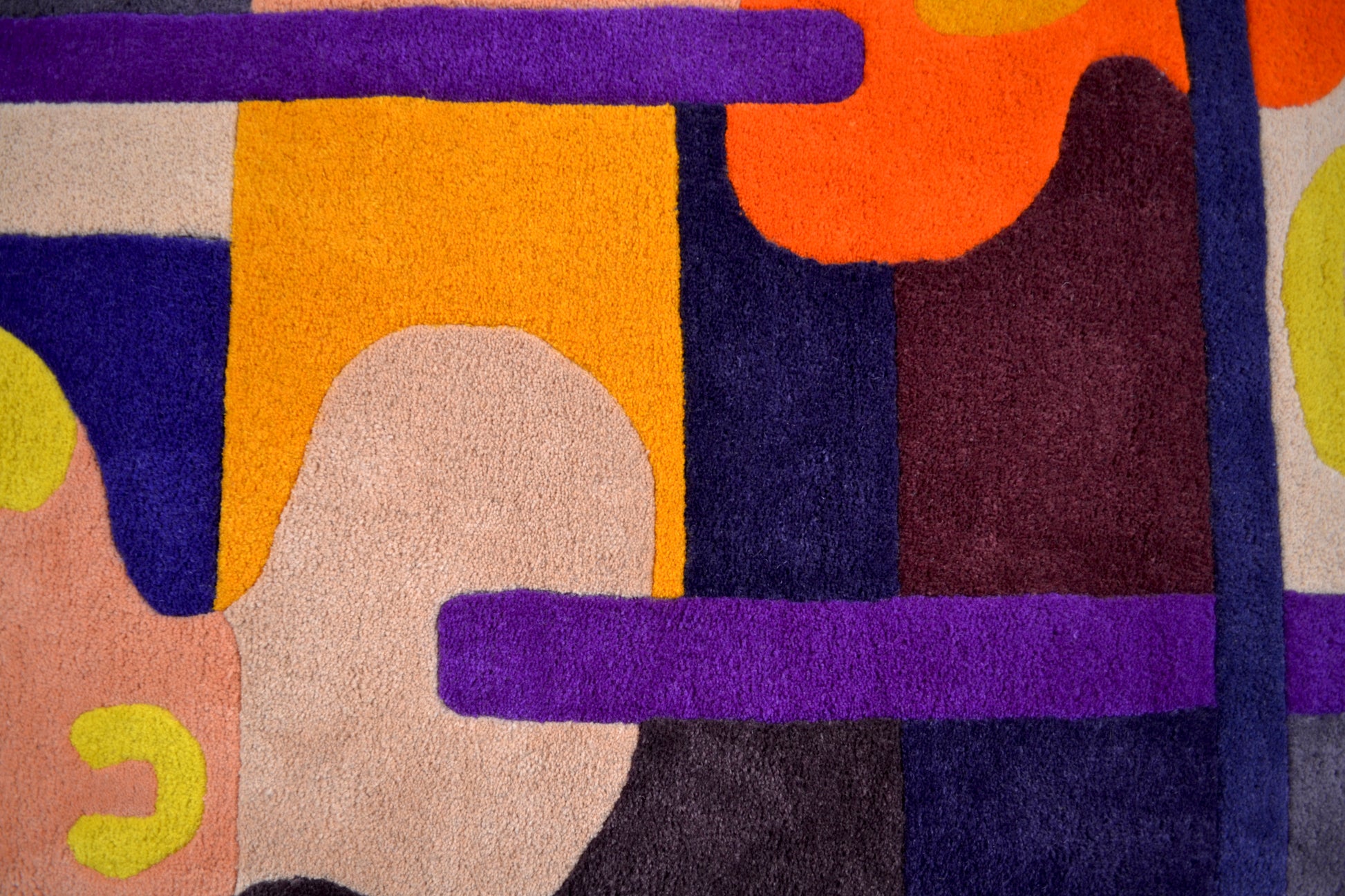 Purple, orange, yellow, cream and burgundy handcrafted tufted wool rug. Abstract shapes and curves. Dark purple border with block colours. Photograph zoomed in on middle section showing block colour details.