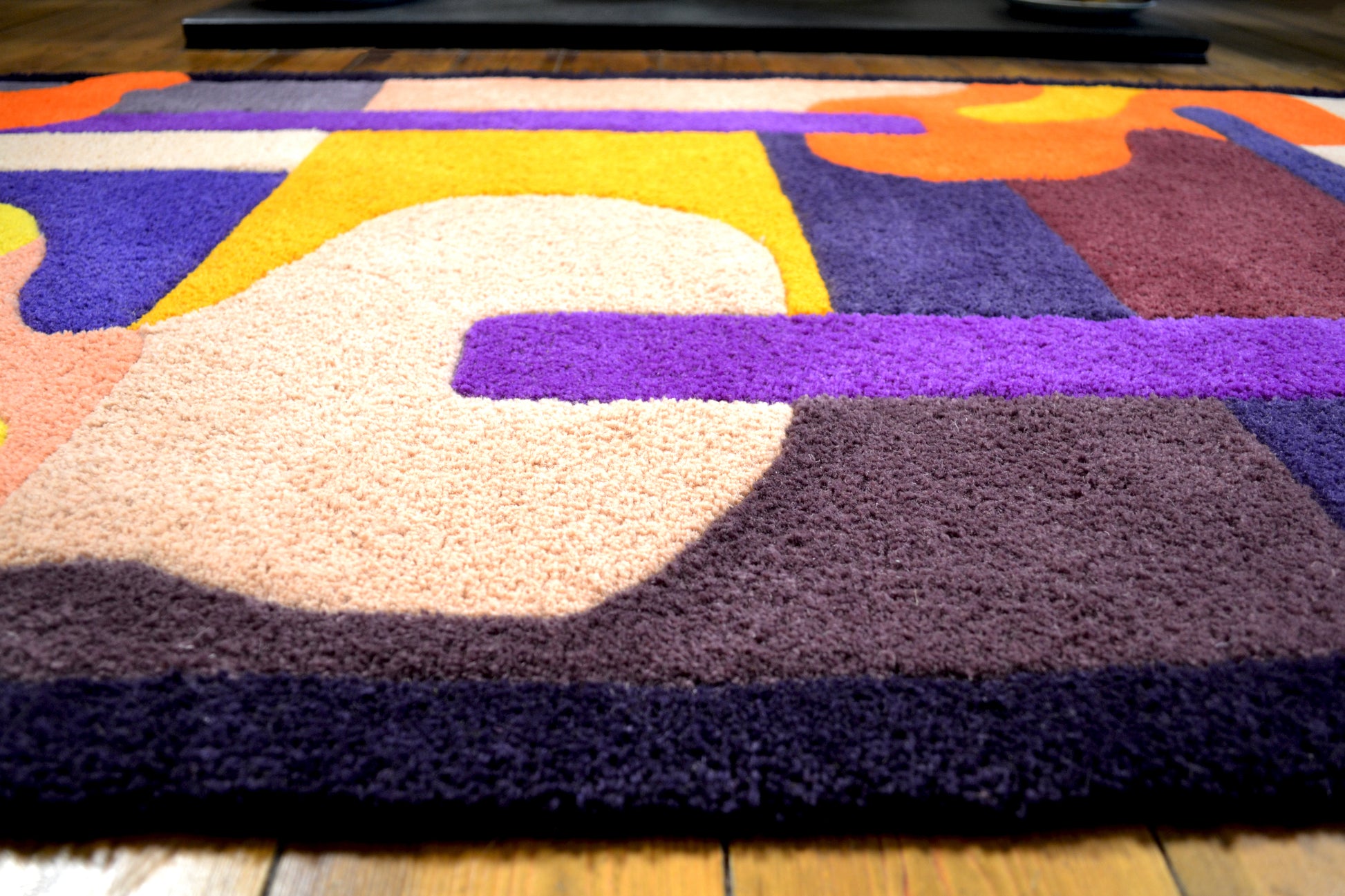 Photograph shows purple, orange, yellow, cream and burgundy handcrafted tufted wool rug. Abstract shapes and curves. Dark purple border with block colours. Pictured on a wooden floorboards. Photographed is a zoomed in middle section showing the thickness of the rug pile.