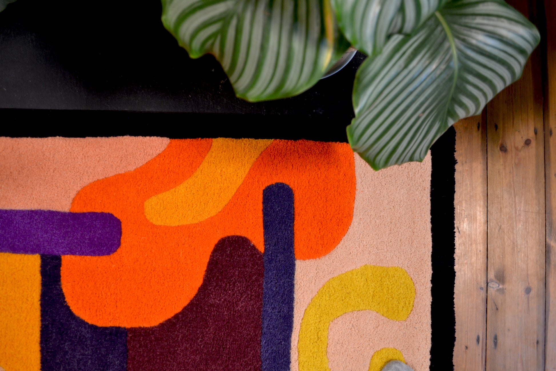 Photographed is a purple, orange, yellow, cream and burgundy handcrafted tufted wool rug. Abstract shapes and curves. Dark purple border with block colours. Pictured on wooden floorboards in front of a fireplace with plant leaning over the rug. Photographed is the upper righthand size corner.