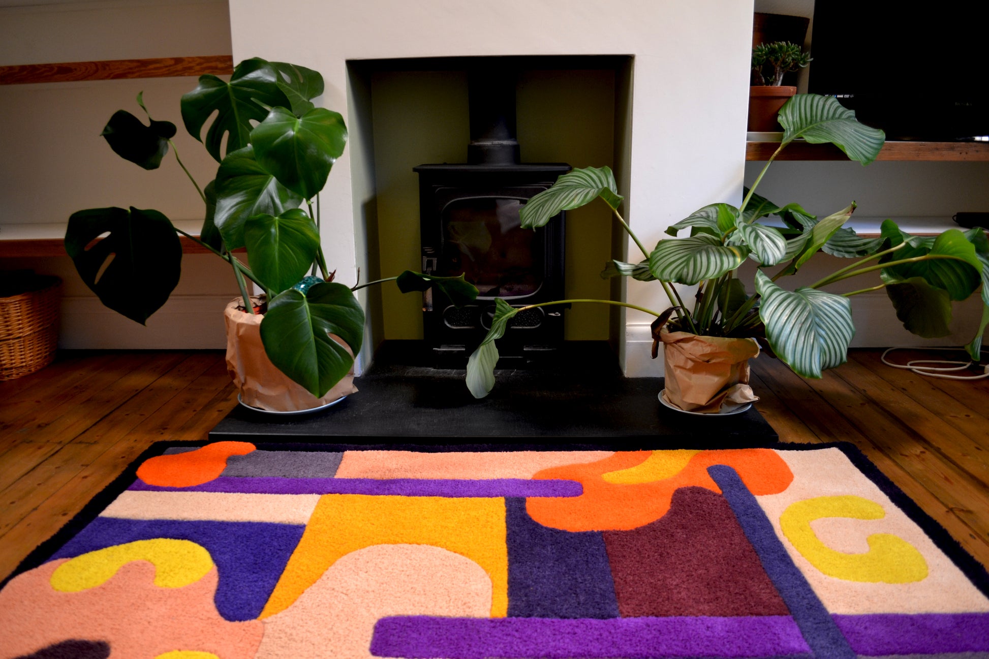 Purple, orange, yellow, cream and burgundy handcrafted tufted wool rug with abstract and curved shapes. Positioned in front of fireplace on stained wooden floorboards in a living room with plants either side of the wood burning fire.