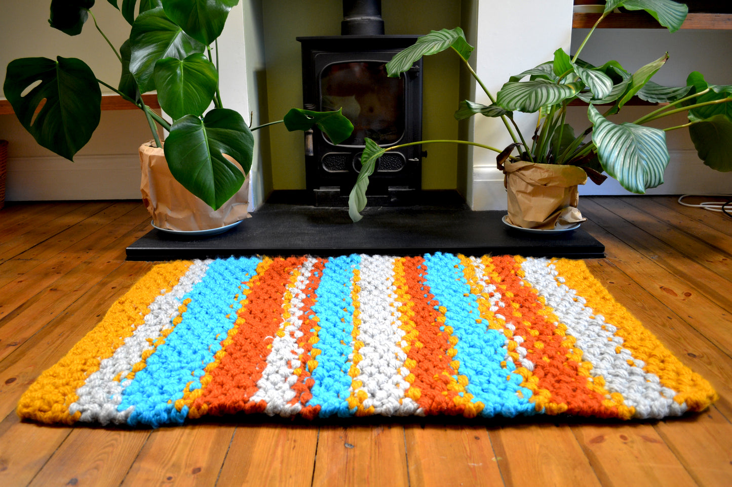 Image shows striped rug in front of wood burner, laying on a wooden floor, with plants placed to the side. Rug is grey, sky blue, burnt orange and mustard yellow striped, with bug mustard yellow stripes at both ends. Rug is laying landscape so the thick pile height of the rug can be seen.