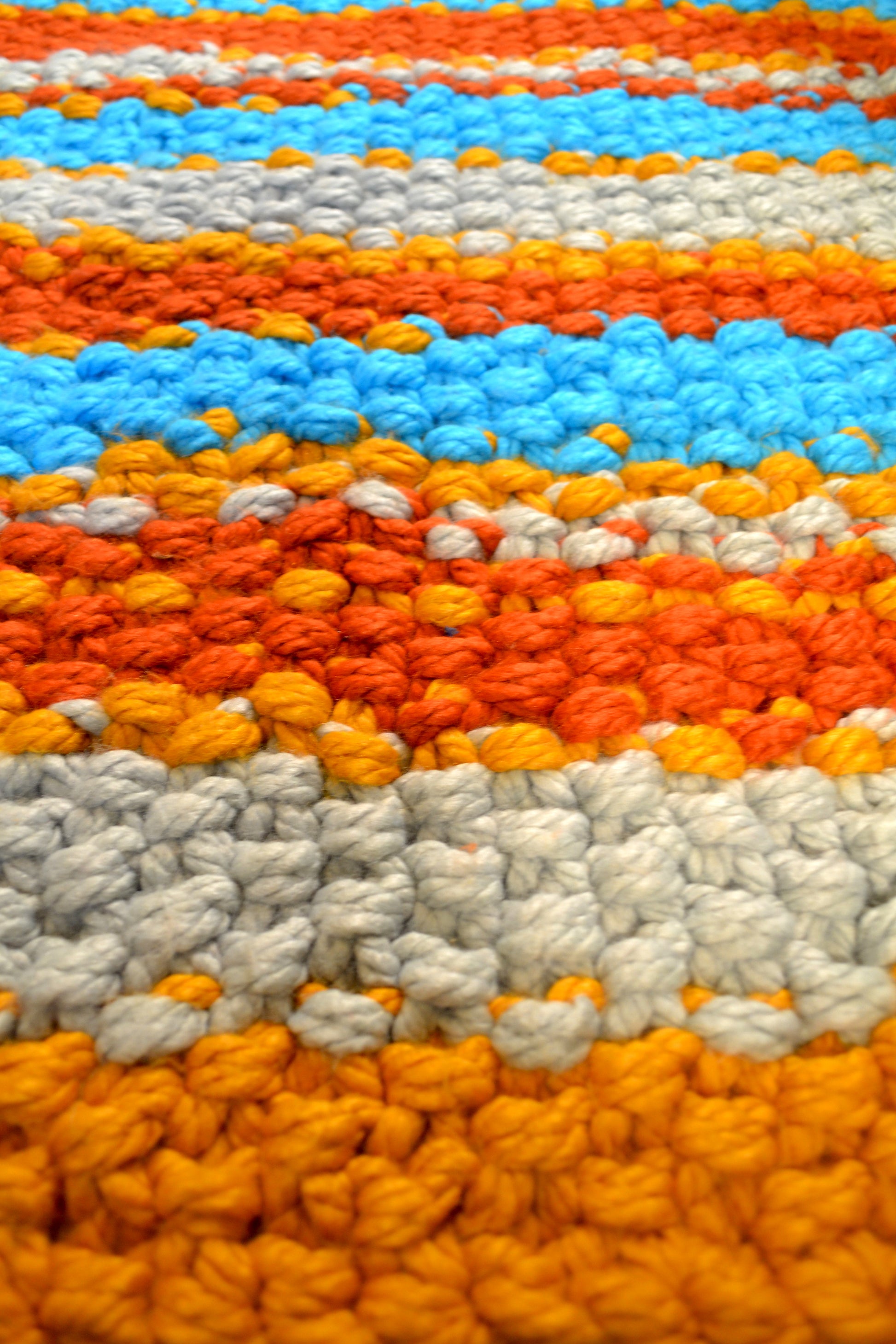 Zoomed in image of striped rug, showing detailing of individual stitches. Rug has a striped pattern. Rug is mustard yellow, burnt orange, grey and crisp sky blue stripes, with thick mustard yellow stripes at both ends. Each individual stitch is visibly and the rug has an extra thick pile height.