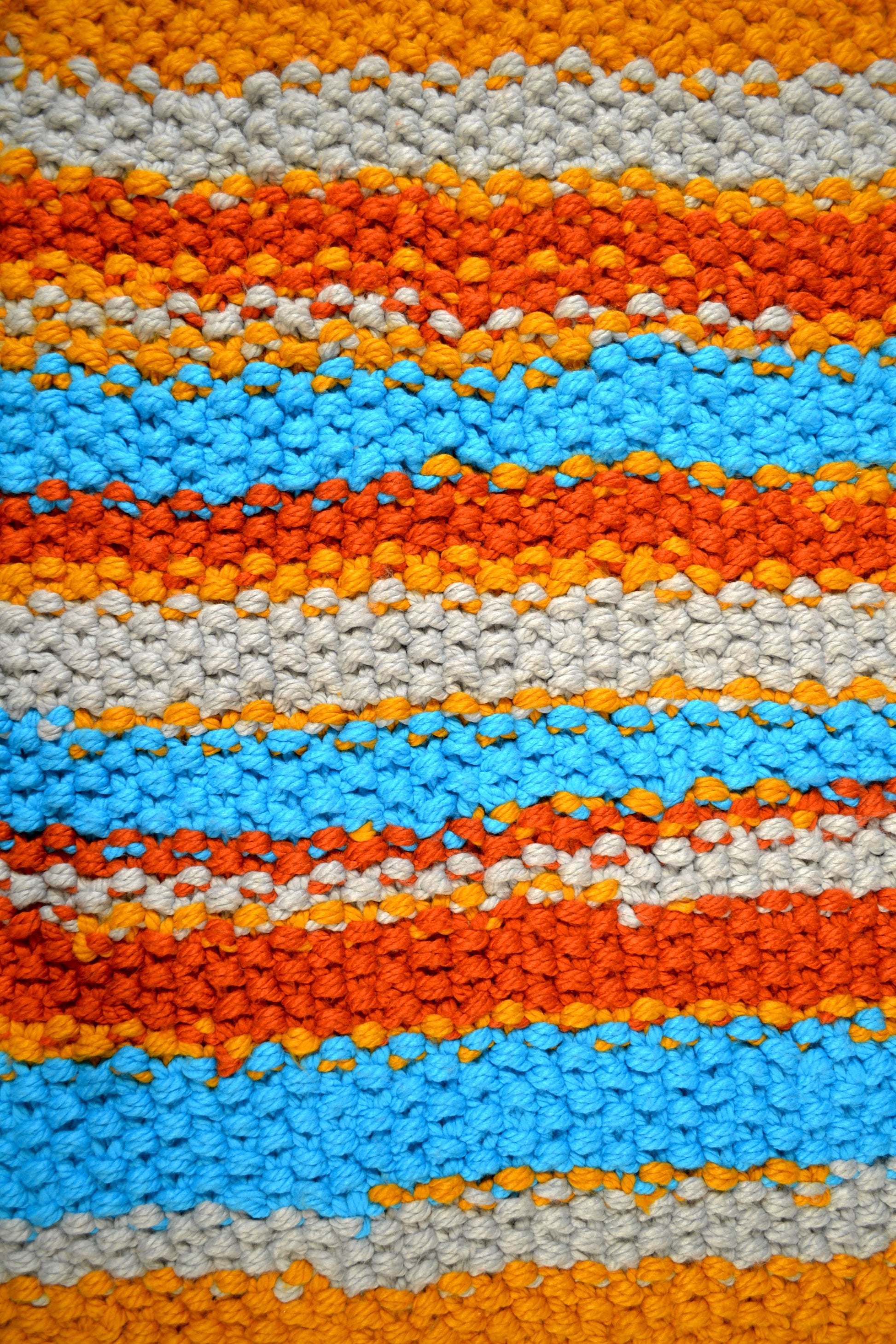 Zoomed in image of striped rug, showing detailing of individual stitches. Rug has a striped pattern. Rug is mustard yellow, burnt orange, grey and crisp sky blue stripes, with thick mustard yellow stripes at both ends. Each individual stitch is visibly and the rug has an extra thick pile height.