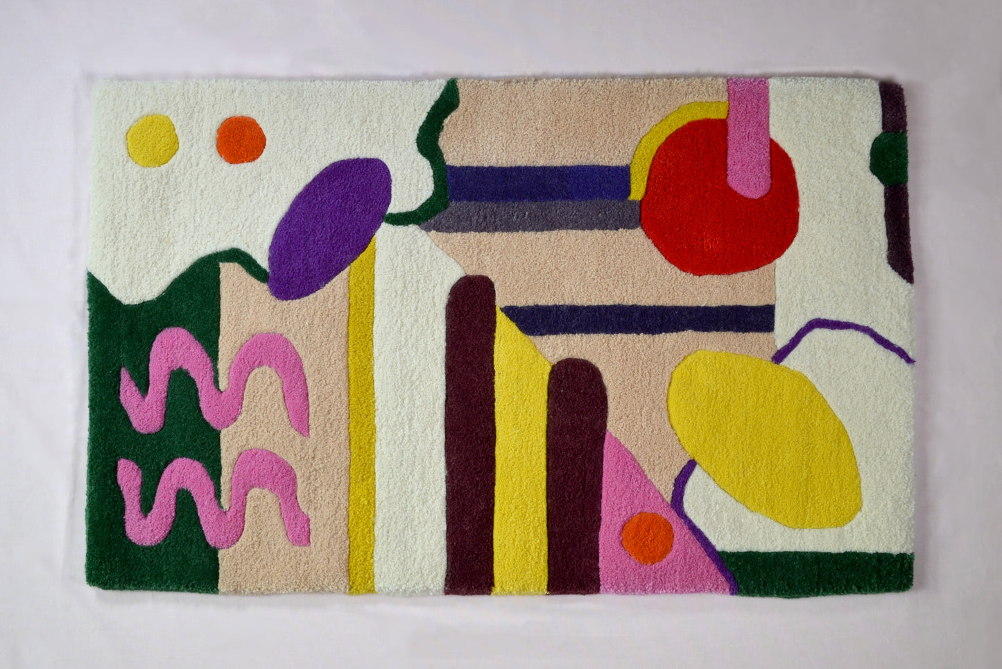 Image shows a cream rug with multicoloured abstract shapes on it. These shapes are red, burgundy, sunflower yellow, hot pink, dark green and orange. the background of the rug has 2 beige block colour sections. The rug is photographed against a very pale pink background.