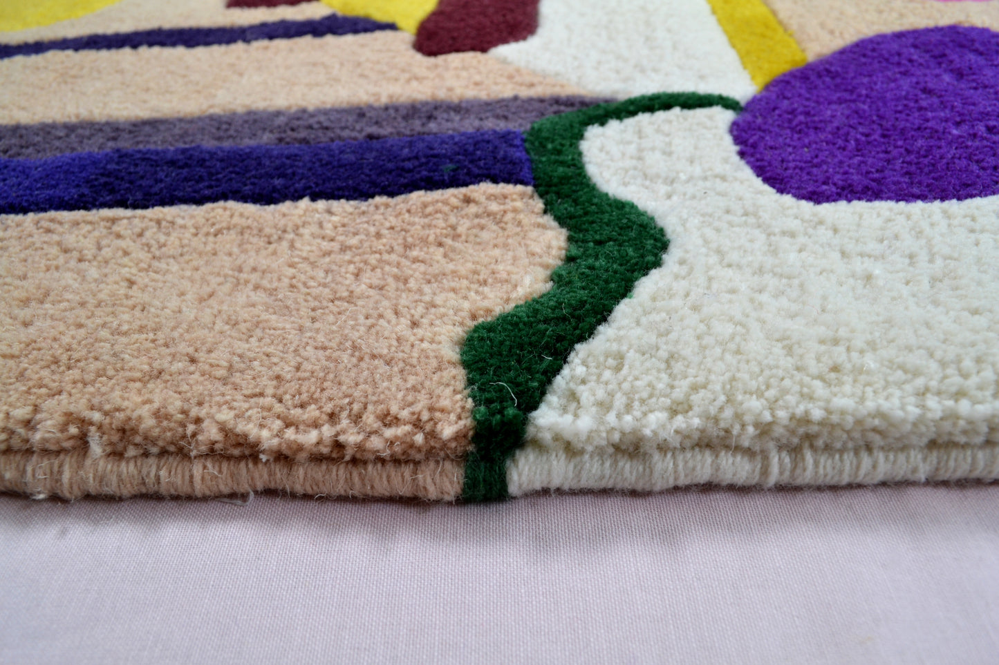 Image shown is a of a small section of the edge of the cream rug. It consists of beige and cream background blocks of colour with a dark green squiggle running through them. Further in the background, just out of focus, can be seen a purple egg shape pattern and a darker purple band of colour. The hand embroidered detailing can be seen along the edge of the rug. The rug is photographed against a very pale pink background.