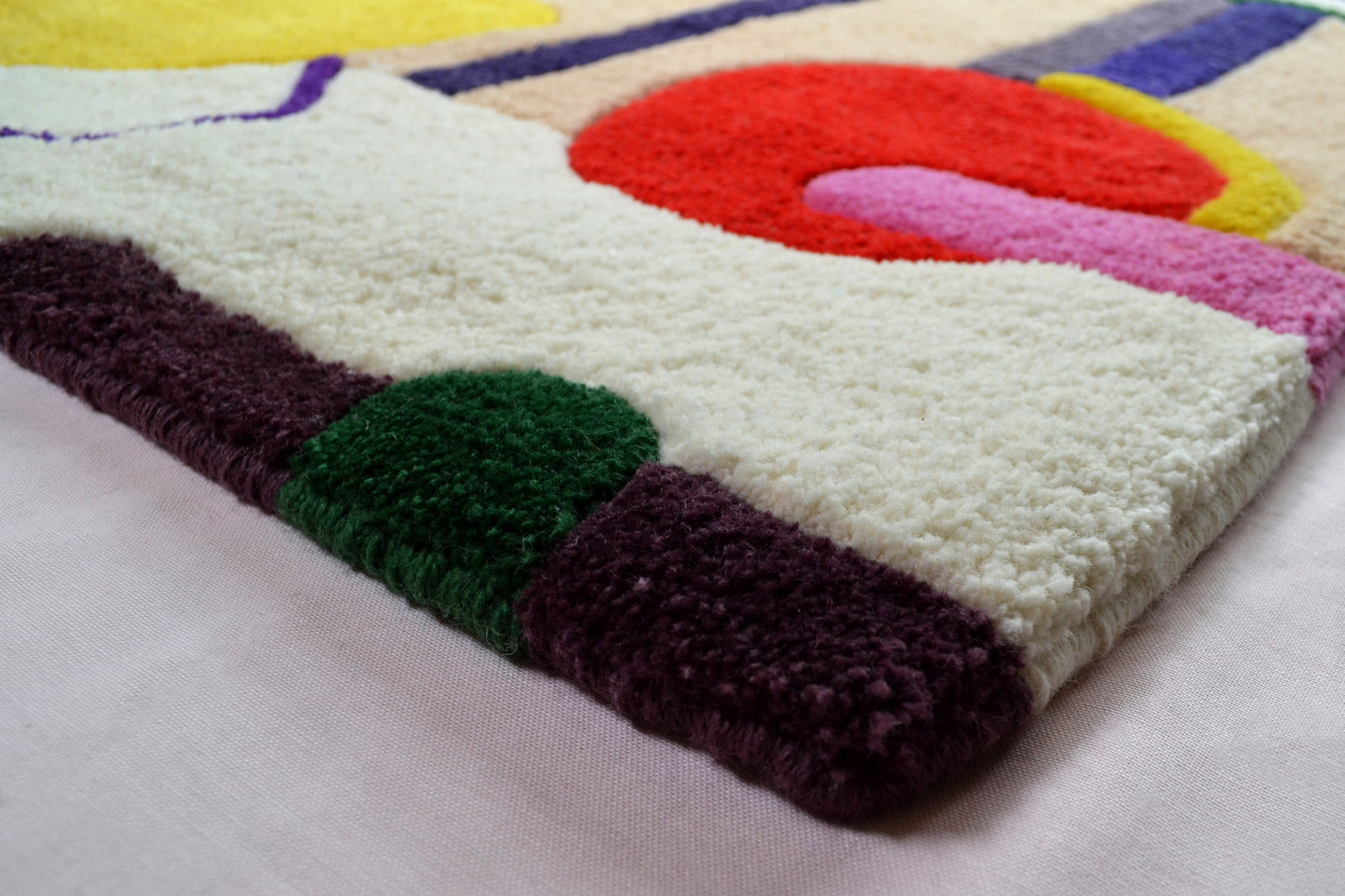 Image shown is of a corner section of the rug showing the hand embroidered detailing along the edge. In the foreground is a dark purple and dark green shapes against the cream background. There can be seen other colourful shapes, however, they are out of focus towards the back of the image. The rug is photographed against a very pale pink background.