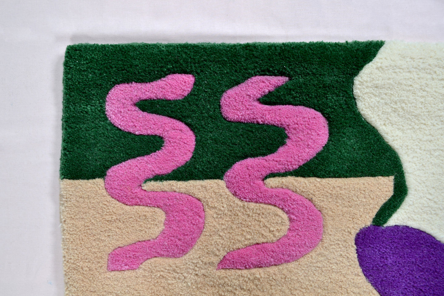 Image consists of a corner section of the rug. The section shown is of dark green and beige block of colour as the background. There is then 2 hot pink curvy squiggle details running across. This corner of the rug is photographed against a very pale pink background.