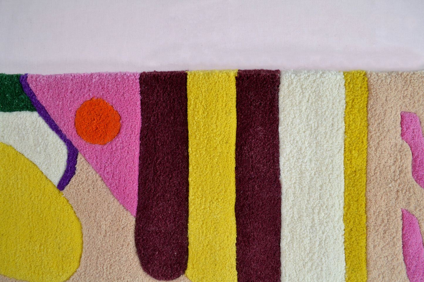 Image shown is of a colourful section of the rug. There can be seen cream, burgundy, beige and yellow bands of colour, with a hot pink triangular section and a small orange circle overlaying that. The rug is photographed against a very pale background.