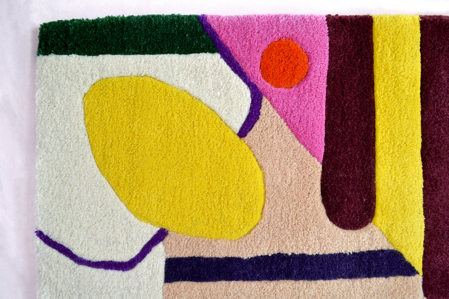 Image is showing a corner section of the cream rug. This consists of a yellow egg shape, with beige, green and cream block section. There is also a small orange circle sitting again a block of hot pink. The corner of this rug is photographed against a very pale pink background.