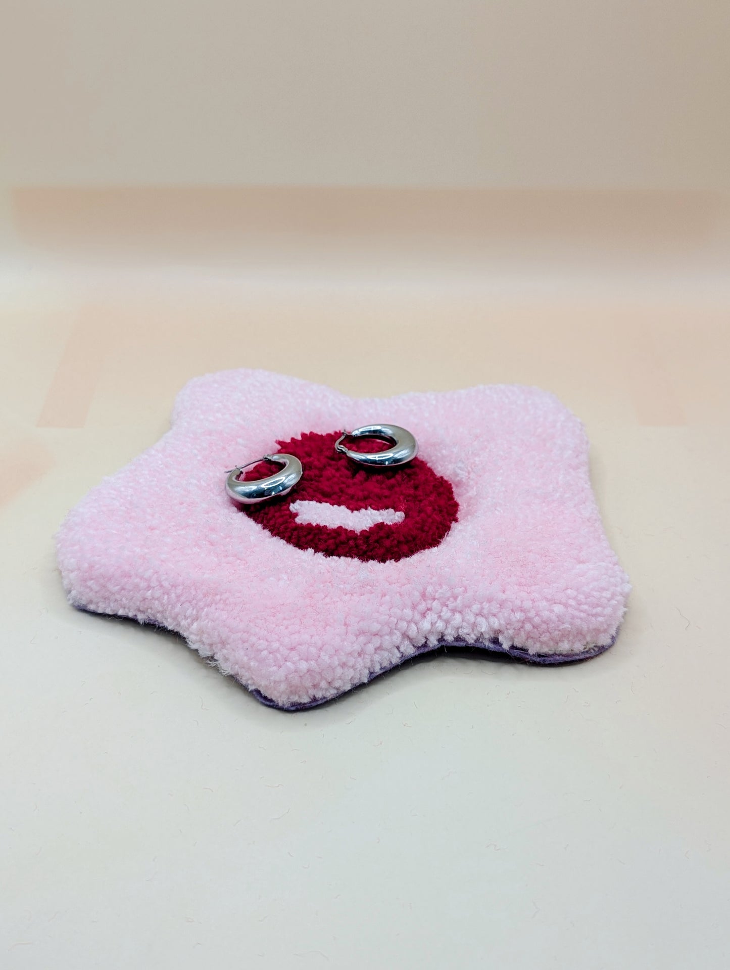 Soft tufted red and pink fried egg shaped mini rug/coaster being used as a jewellery stand, with a small pair of silver hoop earrings resting on it