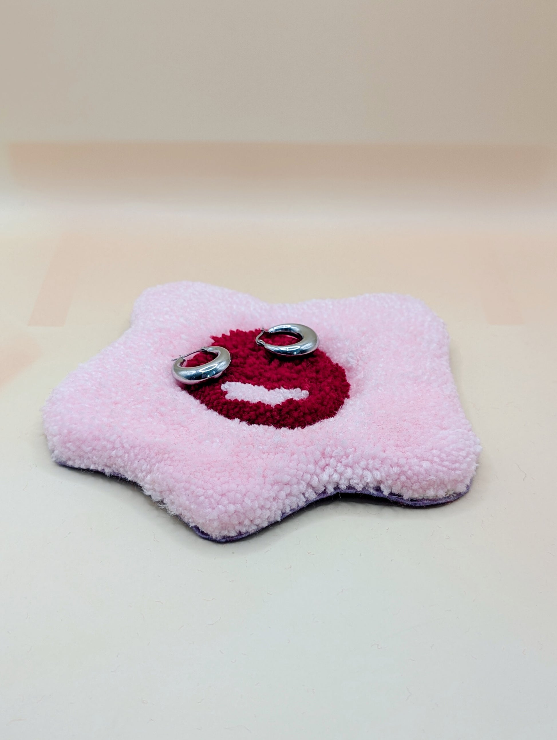 Soft tufted red and pink fried egg shaped mini rug/coaster being used as a jewellery stand, with a small pair of silver hoop earrings resting on it