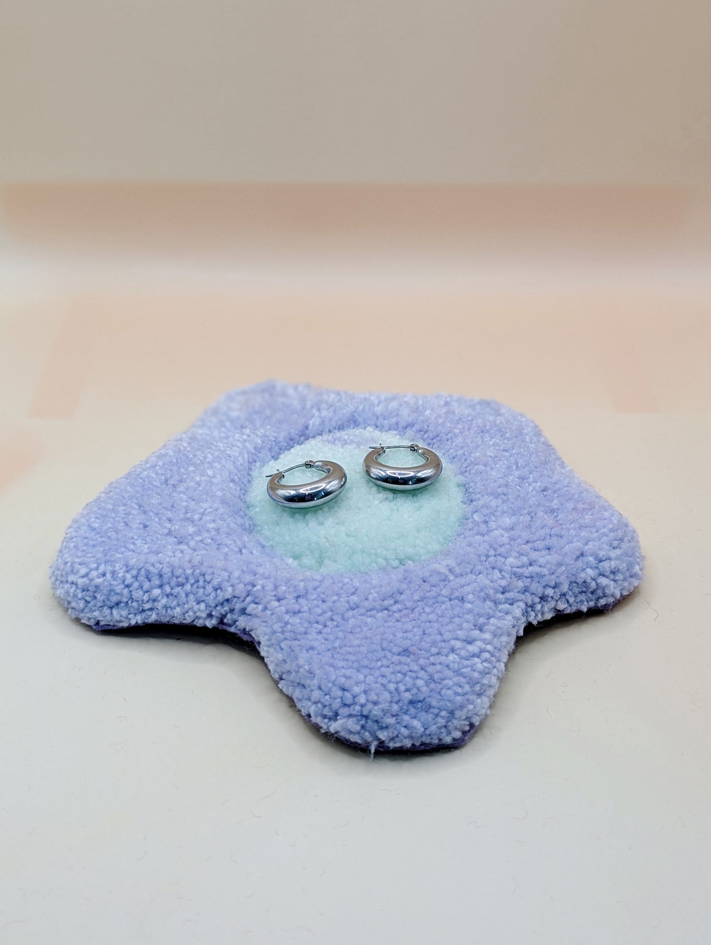 Soft tufted purple and green fried egg shaped mini rug/coaster being used as a jewellery stand, with a small pair of silver hoop earrings resting on it 
