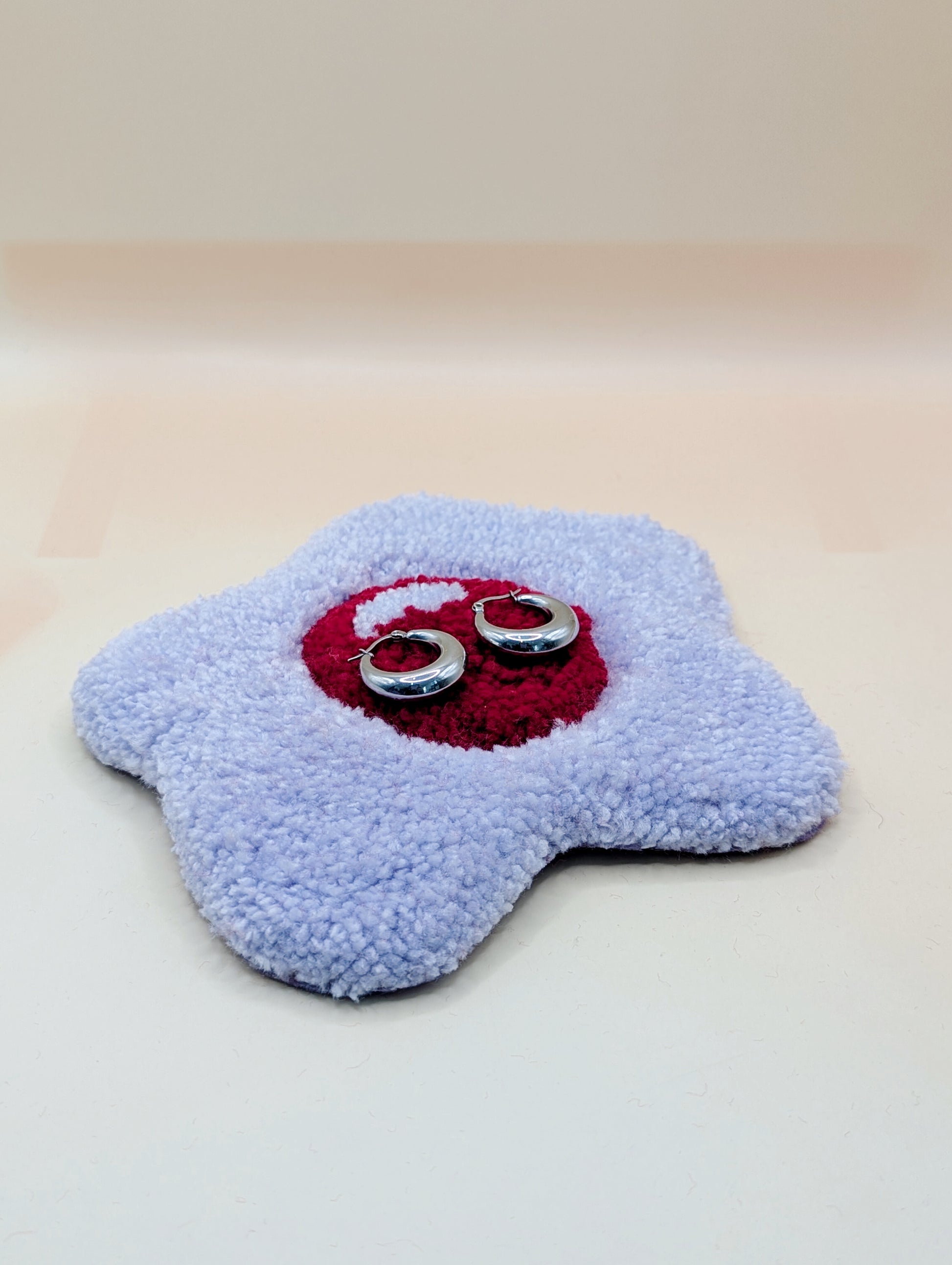 Soft tufted red and pink fried egg shaped mini rug/coaster being used as a jewellery stand, with a pair of small silver hoops resting on it