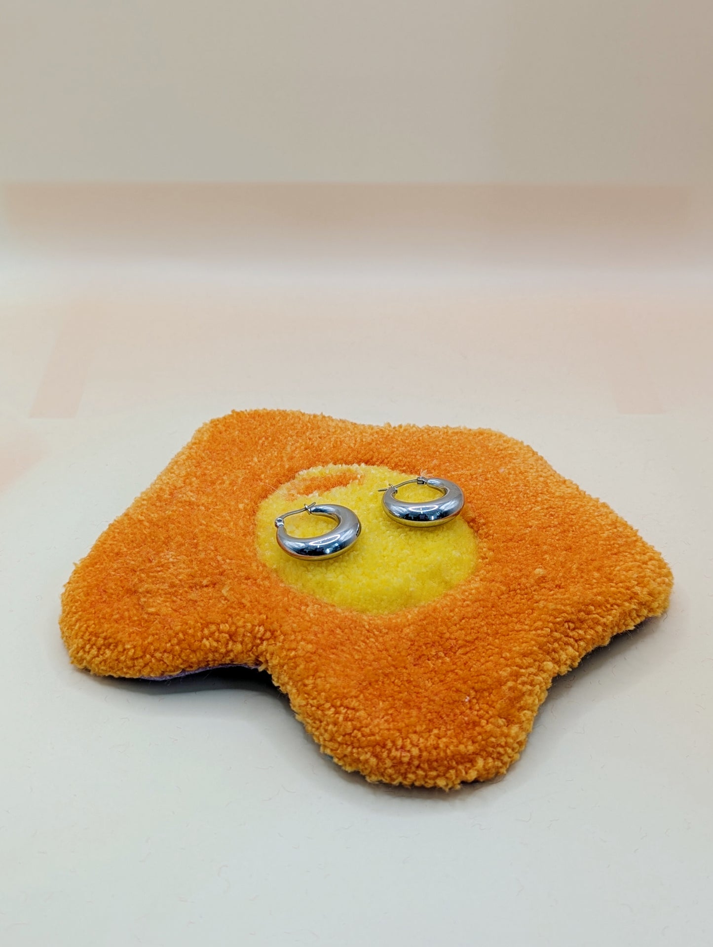 Soft tufted red and pink fried egg shaped mini rug/coaster being used a jewellery stand, with a small pair of silver hoop earrings resting on it 
