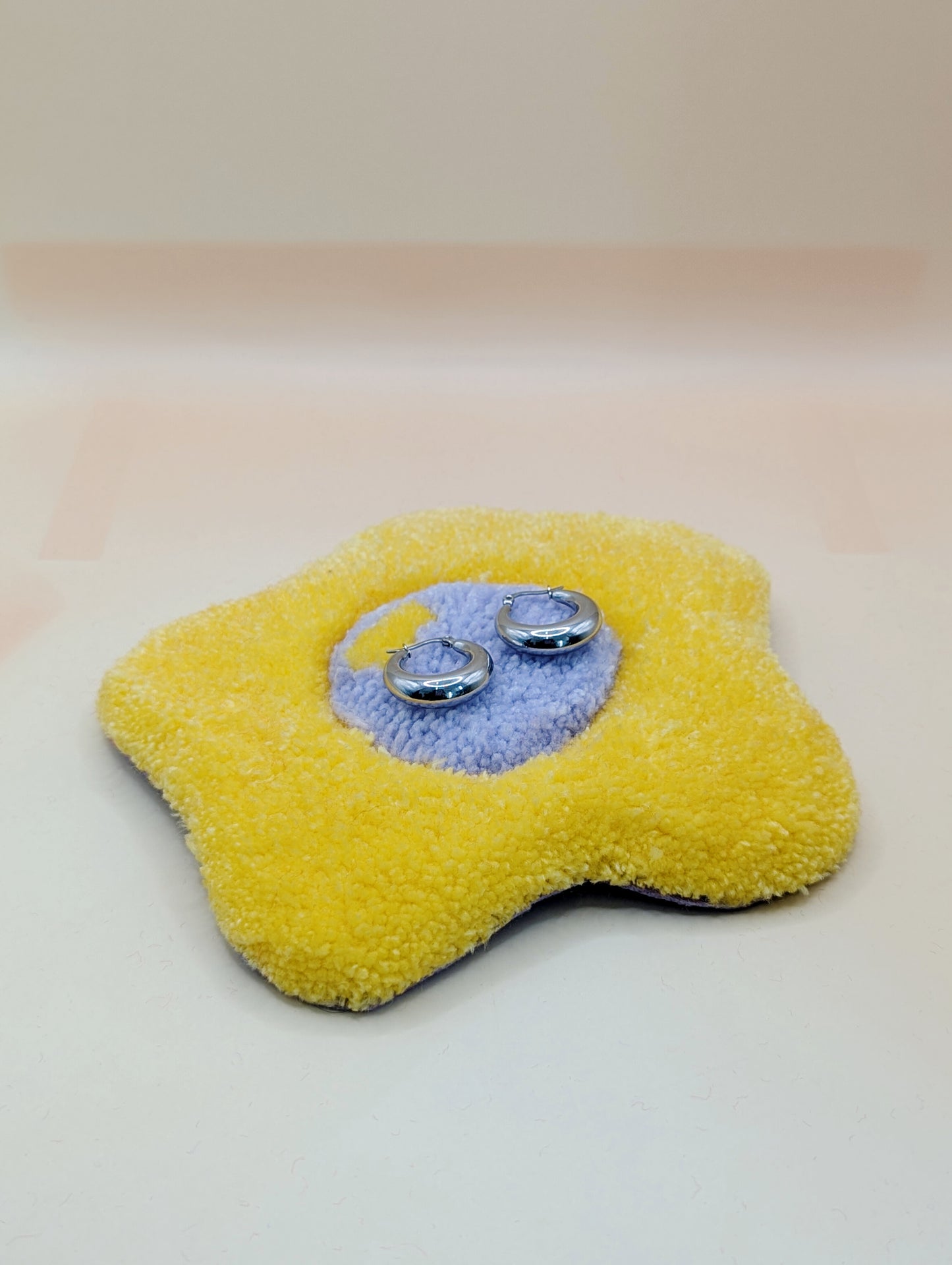 Soft tufted yellow and purple fried egg shaped mini rug/coaster being used as a jewellery stand, with a small pair of silver hoops resting on it 