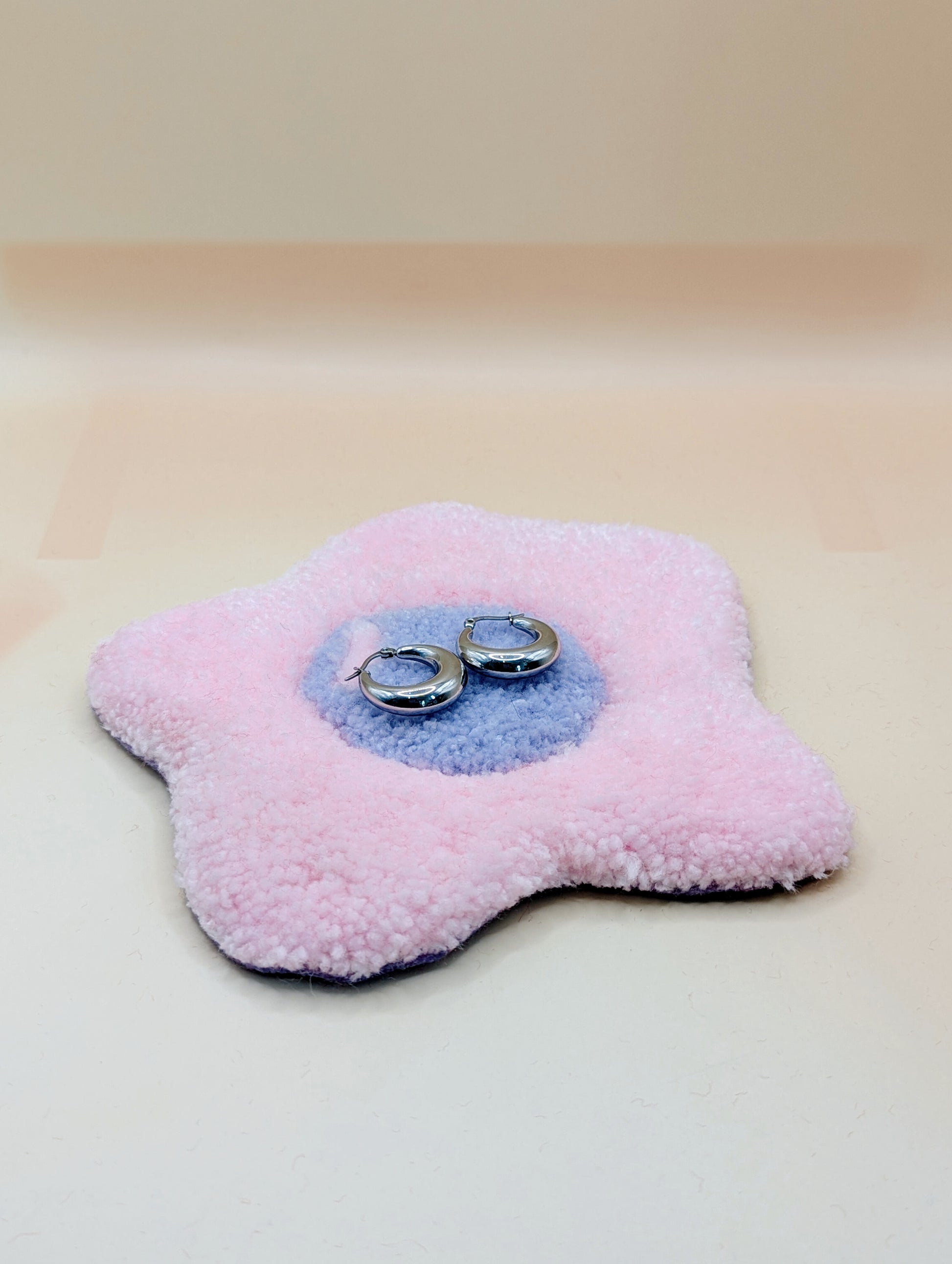 Soft tufted pink and purple fried egg shaped mini rug/coaster being used as a jewellery stand, with a small pair of silver hoop earrings resting on it