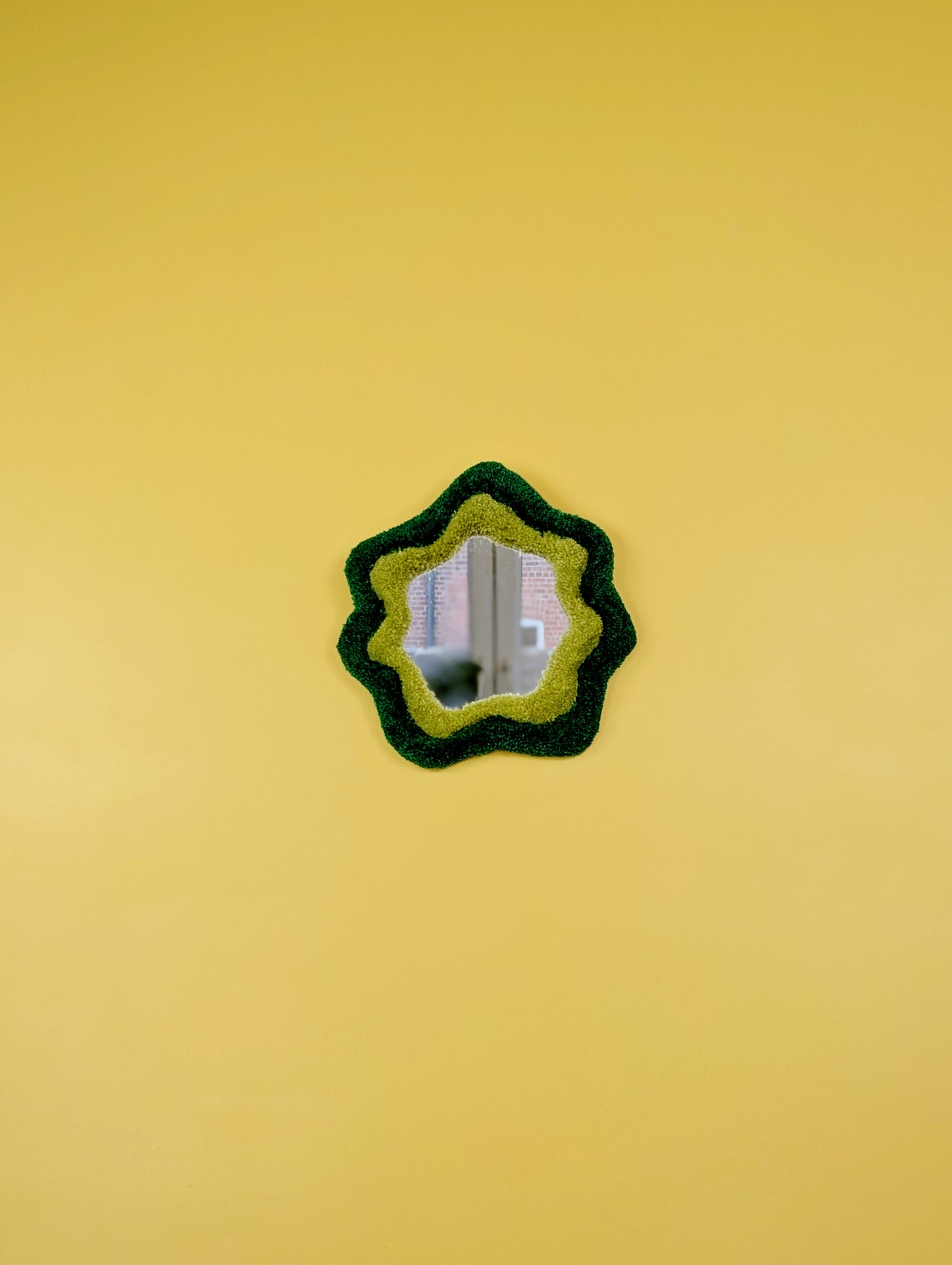  Handmade green tufted mirror with organic wavy shape hanging on a yellow wall.