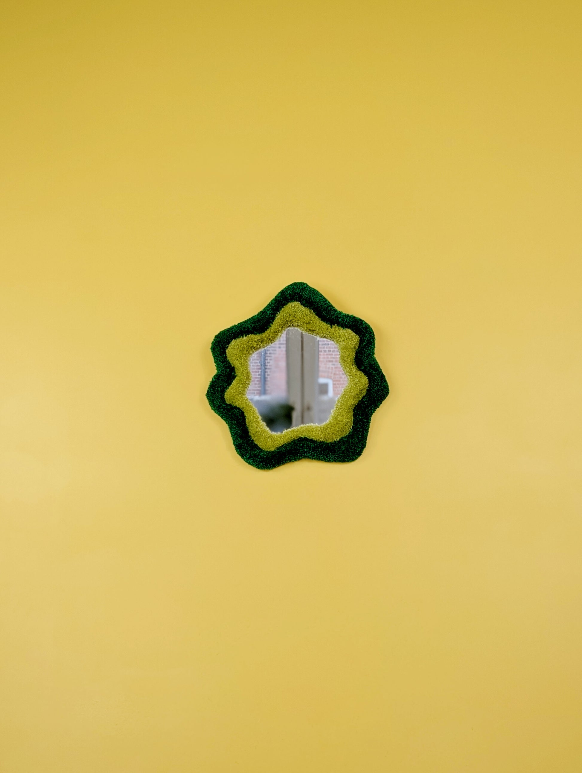  Handmade green tufted mirror with organic wavy shape hanging on a yellow wall.