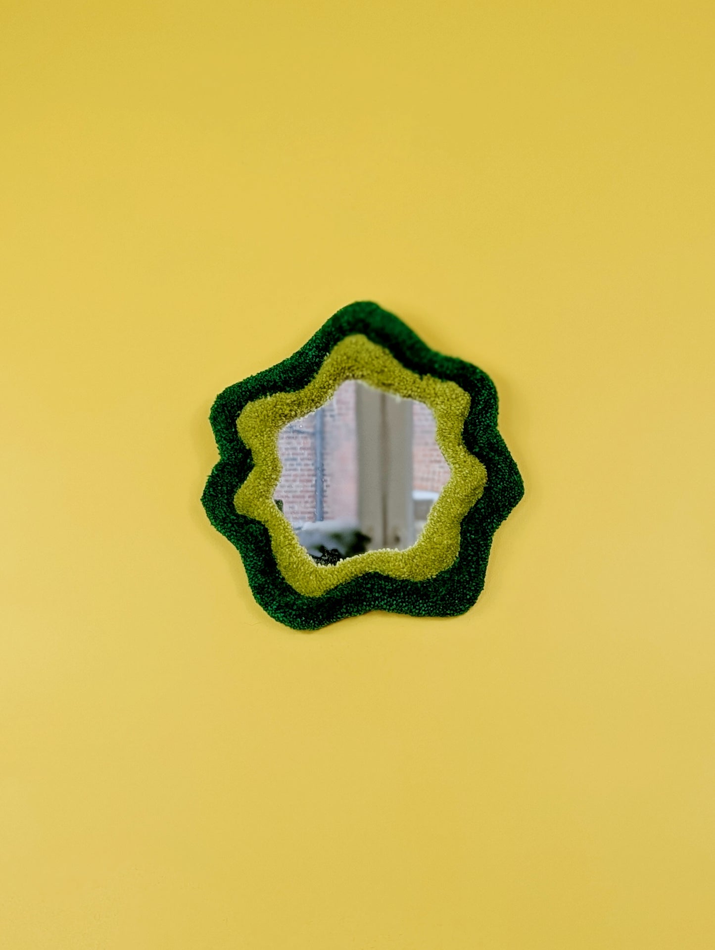 Handmade green tufted mirror with organic wavy shape hanging on a yellow wall.