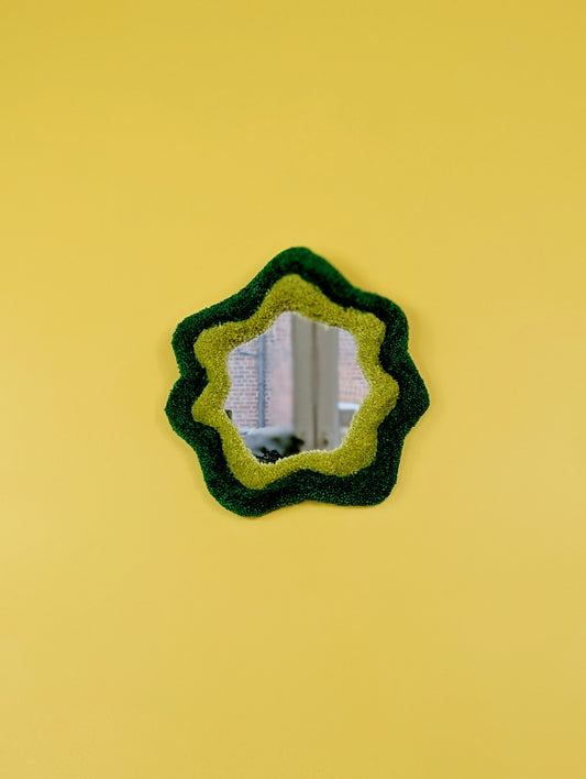 Handmade green tufted mirror with organic wavy shape hanging on a yellow wall.