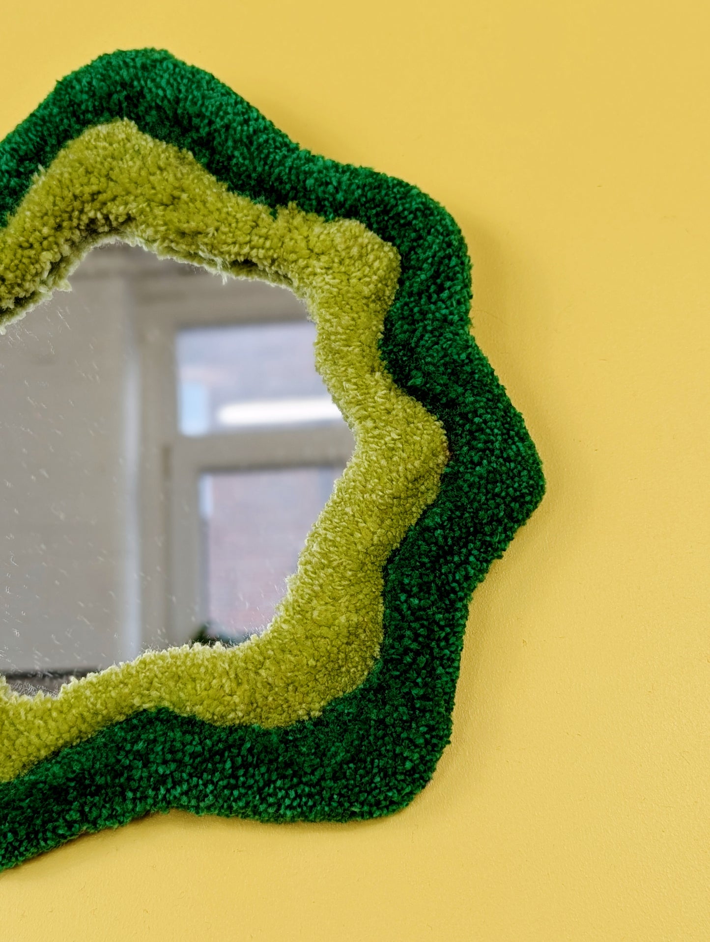 Handmade green tufted mirror with organic wavy shape hanging on a yellow wall.