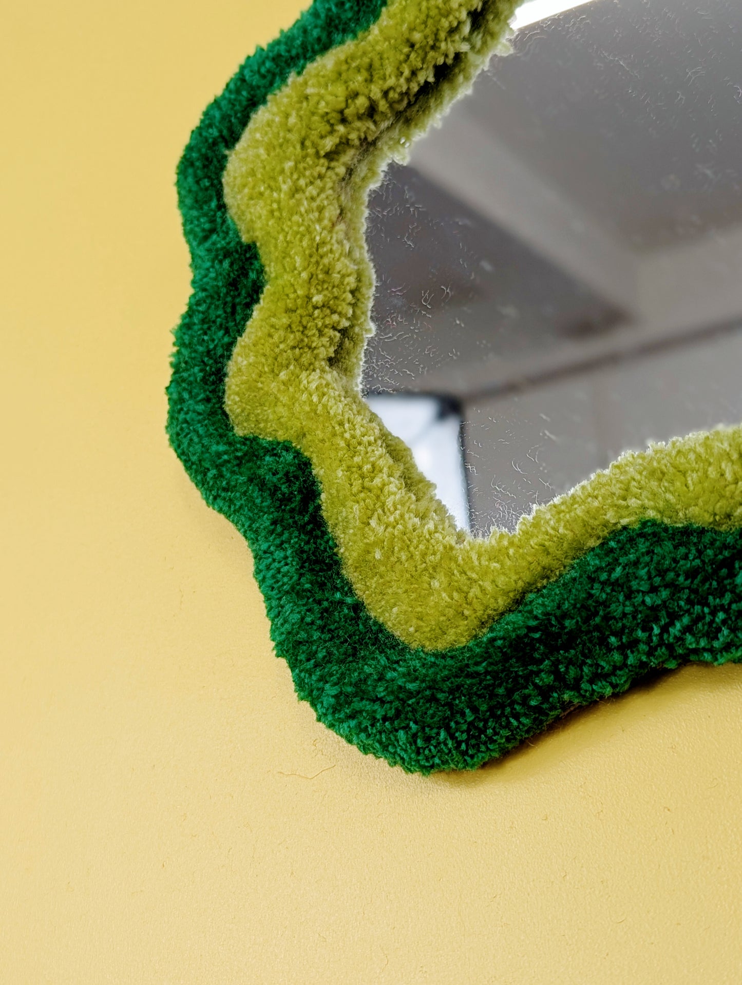 Handmade green tufted mirror with organic wavy shape hanging on a yellow wall.