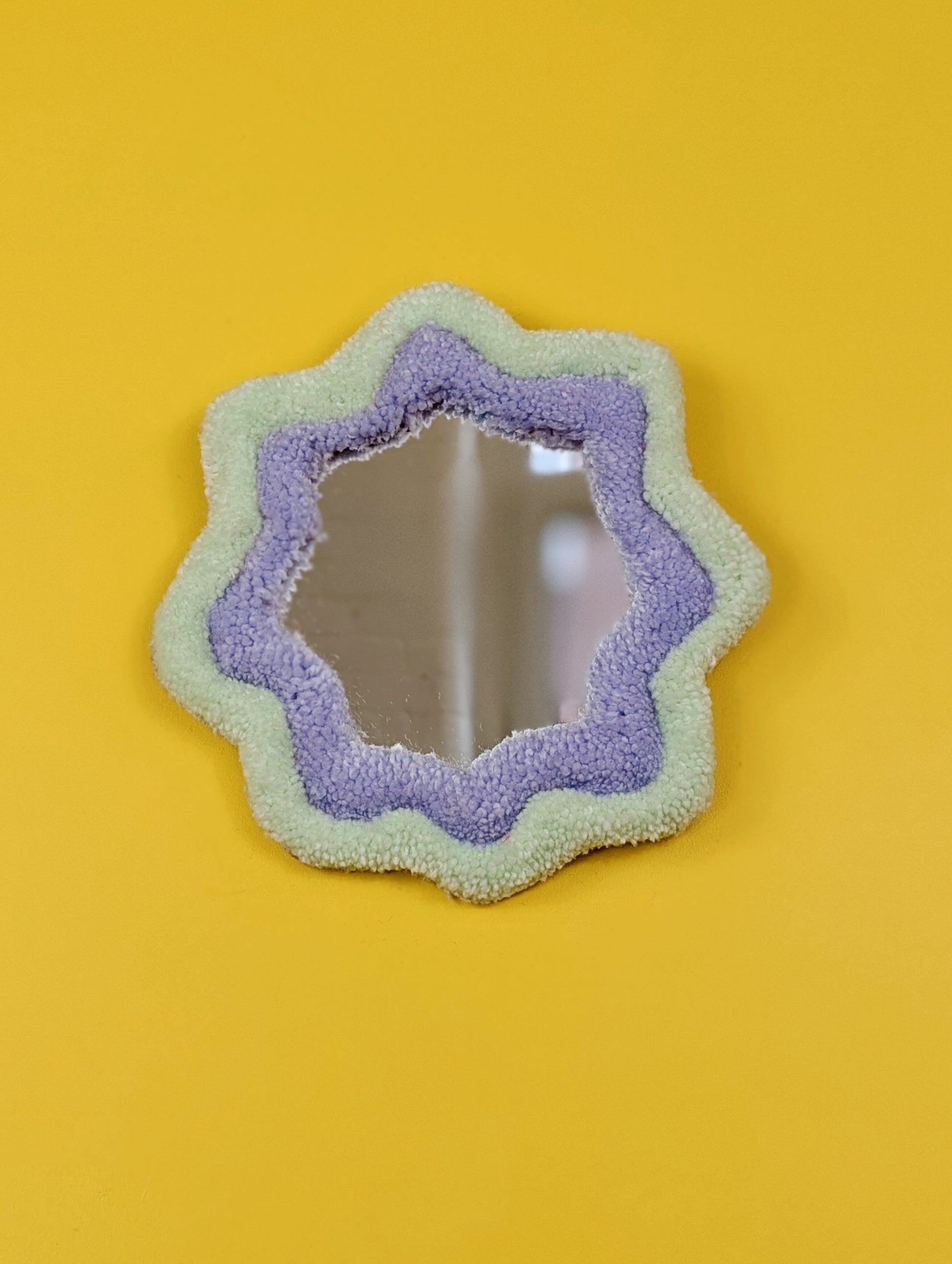 Handmade pale green and purple tufted mirror with organic wavy shape hanging on a yellow wall