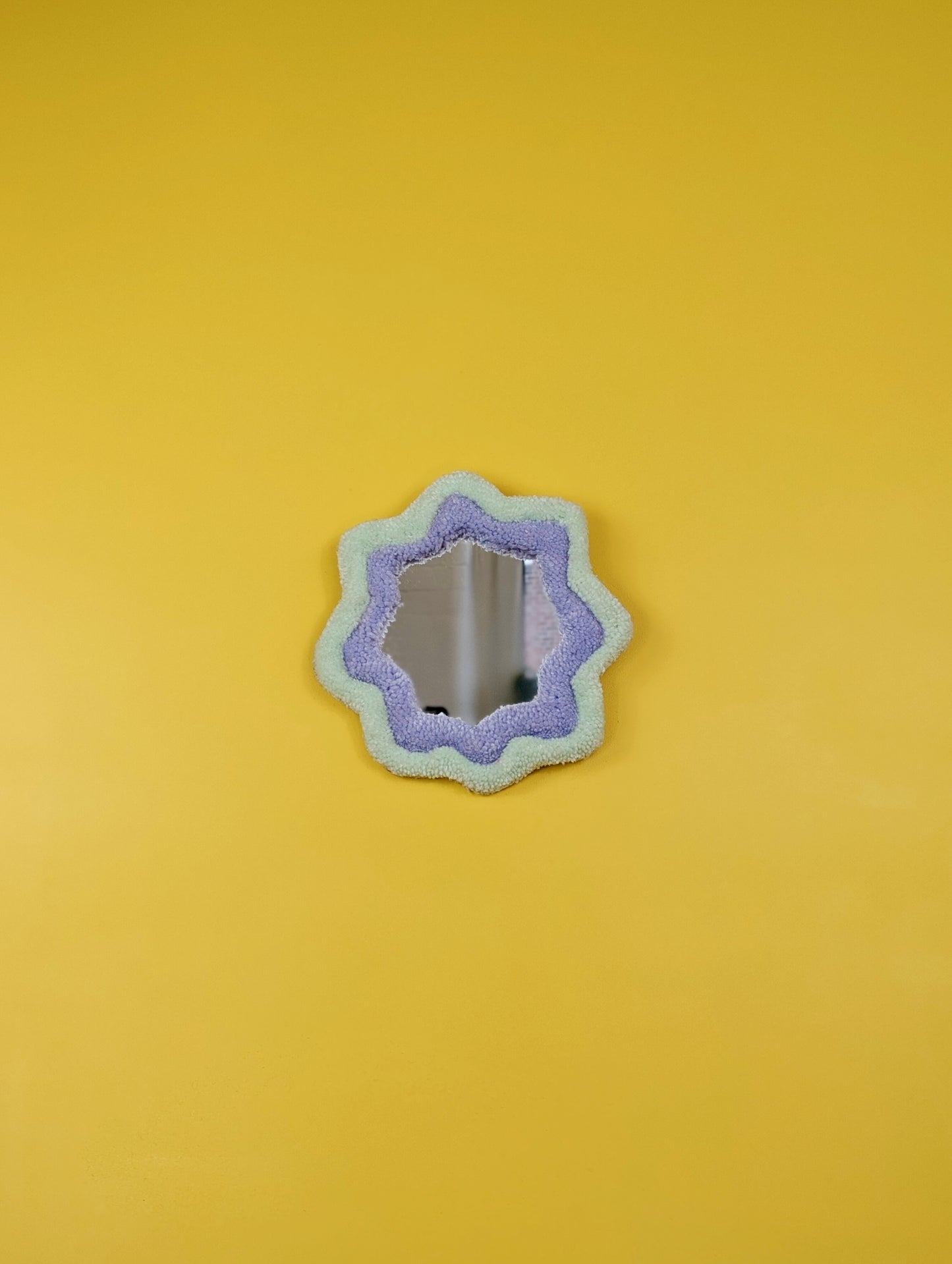 Handmade pale green and purple tufted mirror with organic wavy shape hanging on a yellow wall