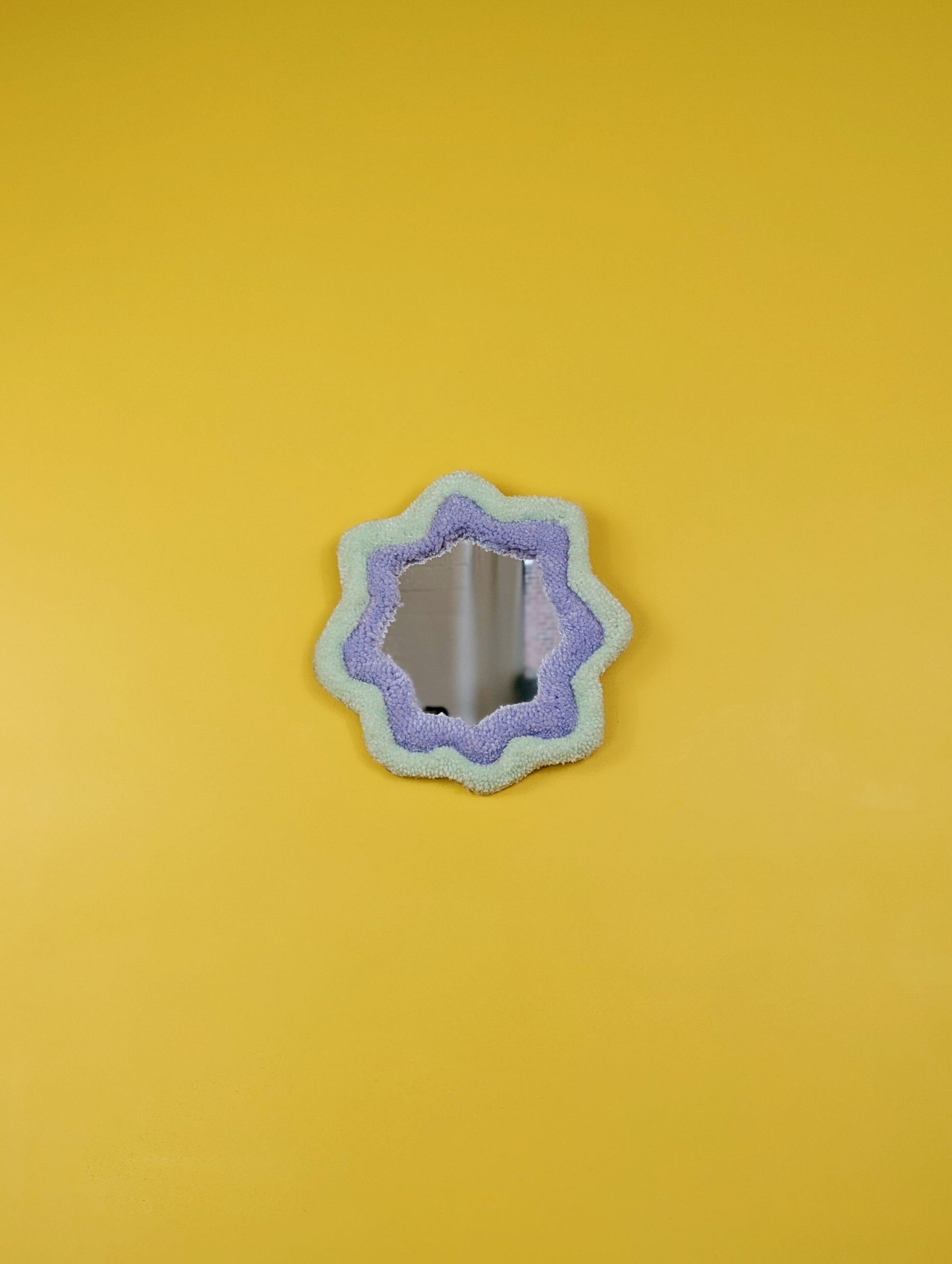 Handmade pale green and purple tufted mirror with organic wavy shape hanging on a yellow wall