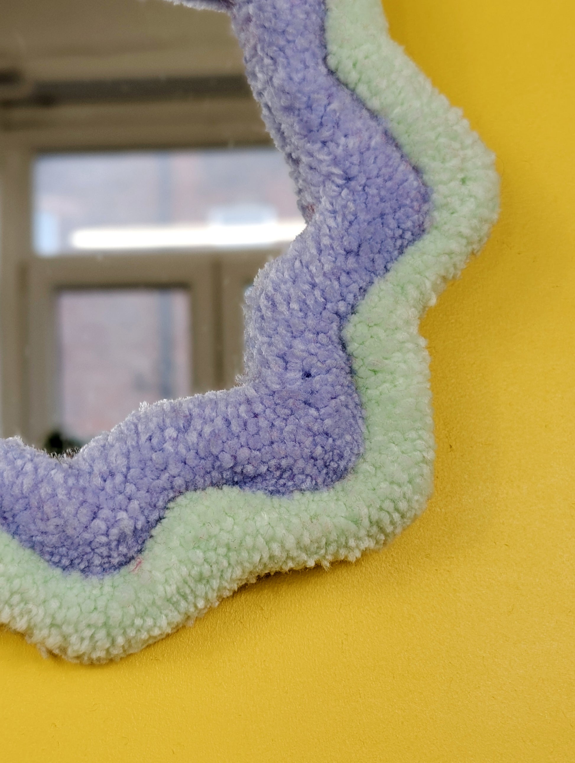 Handmade pale green and purple tufted mirror with organic wavy shape hanging on a yellow wall