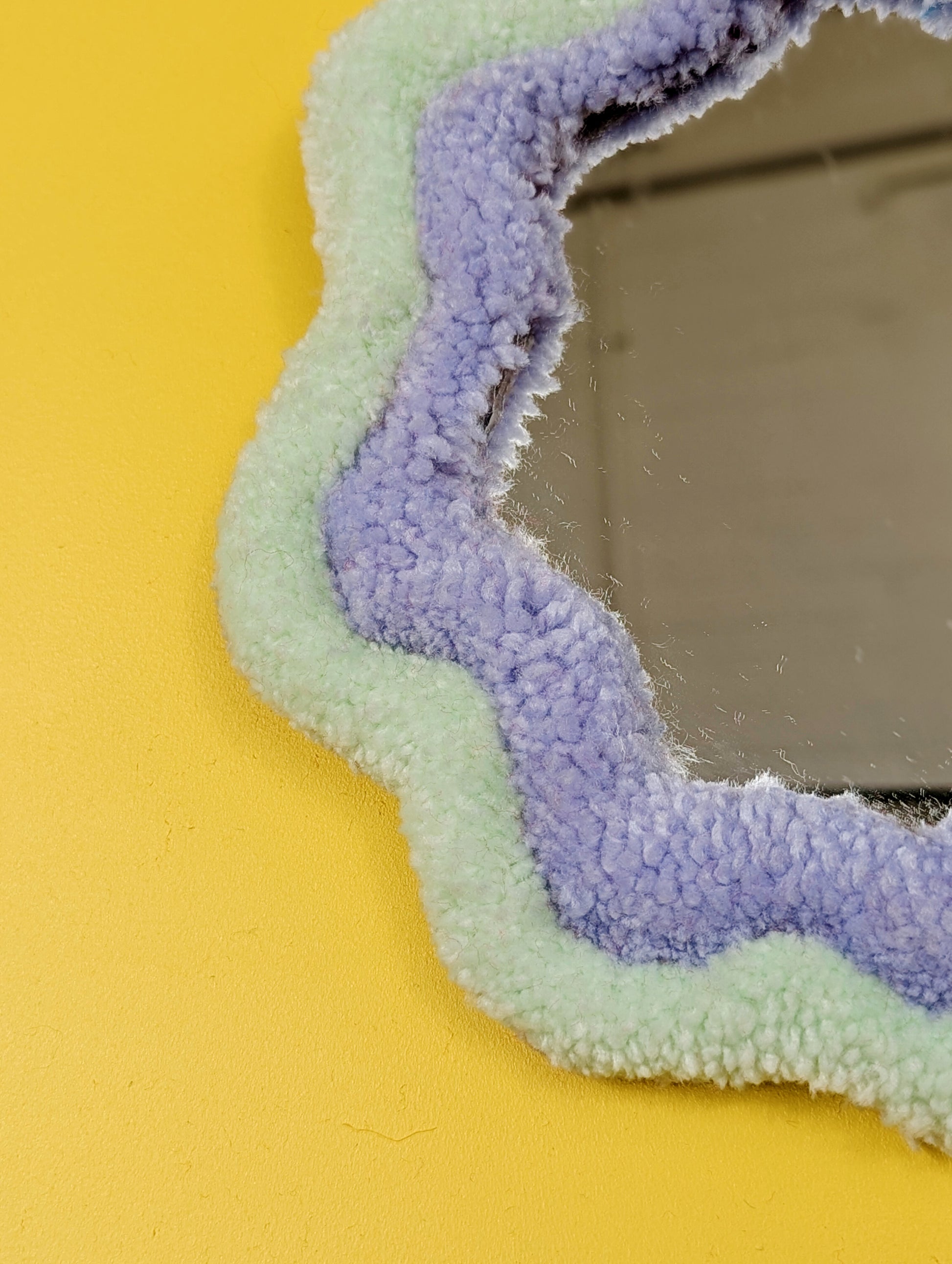 Handmade pale green and purple tufted mirror with organic wavy shape hanging on a yellow wall