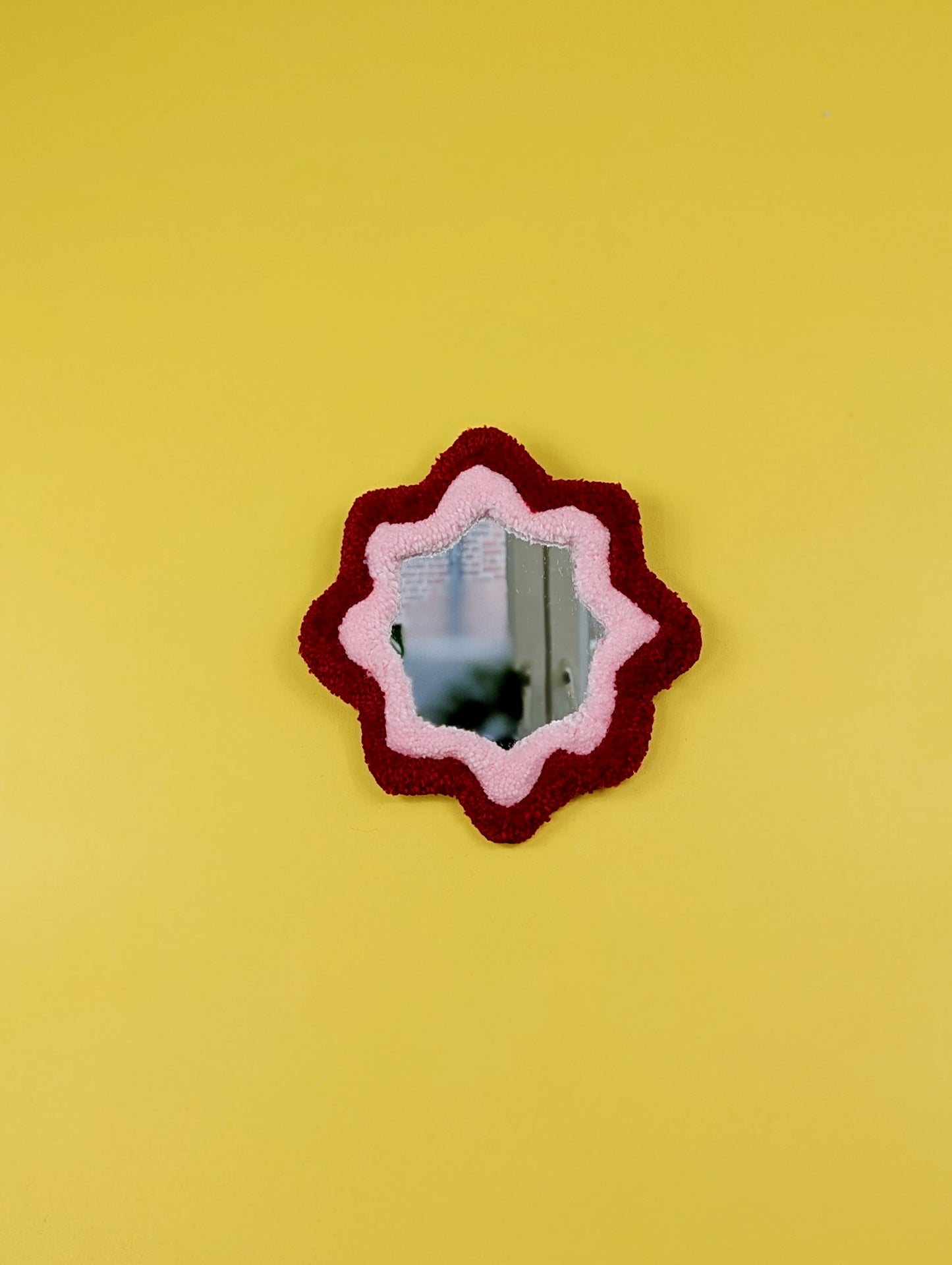  Handmade red and pink tufted mirror with organic wavy shape hanging on a yellow wall.