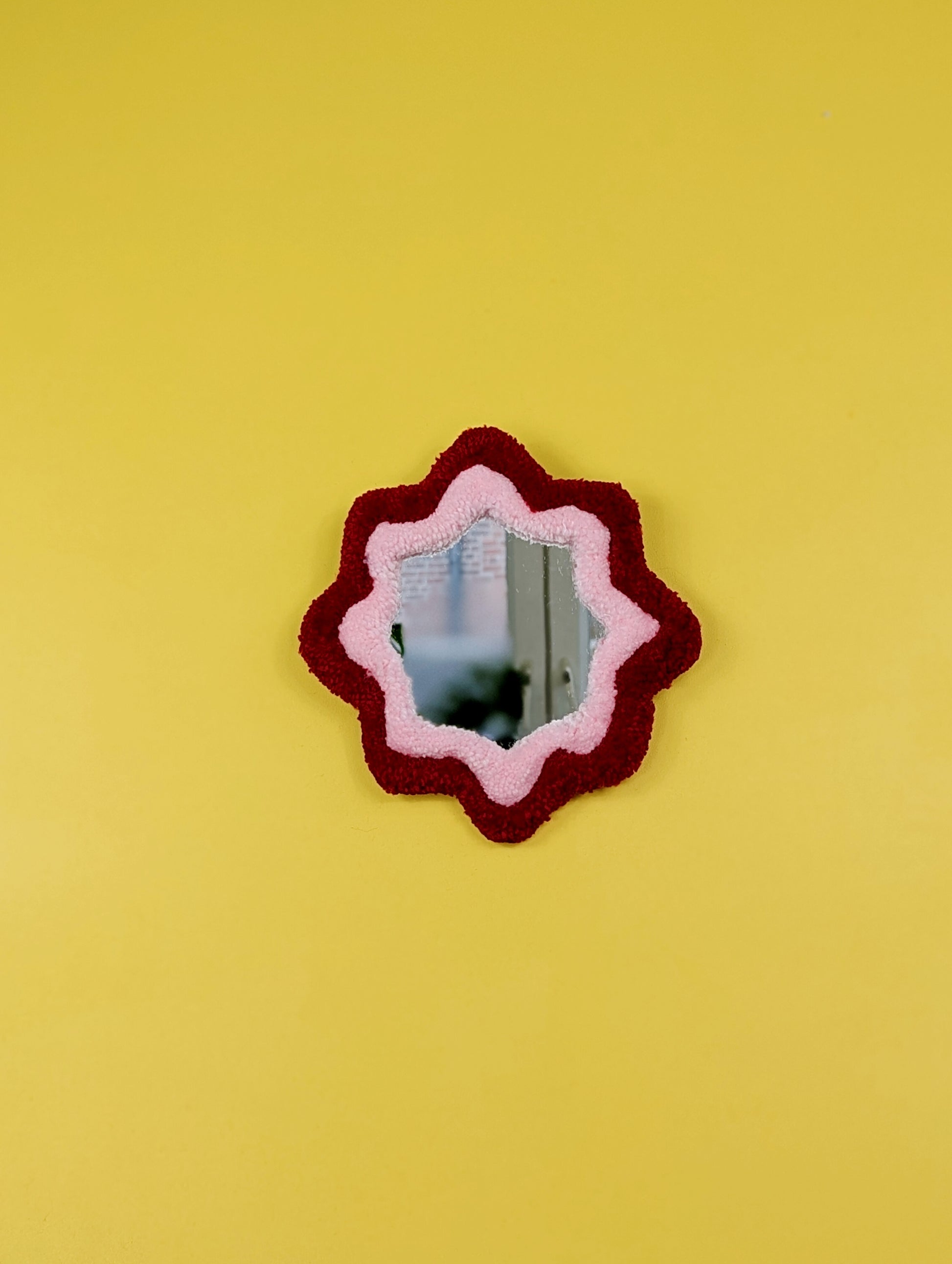  Handmade red and pink tufted mirror with organic wavy shape hanging on a yellow wall.