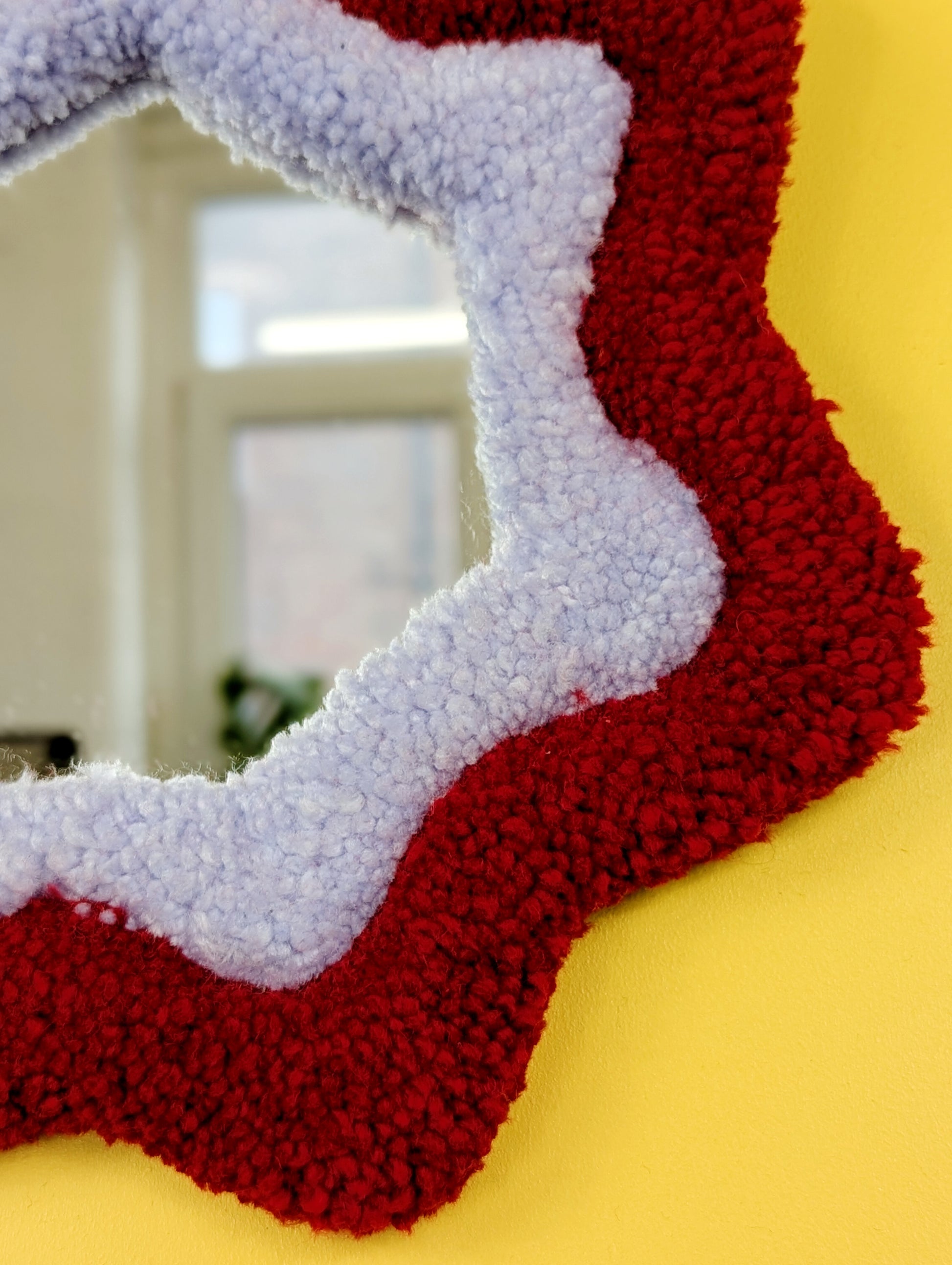  Handmade red and purple tufted mirror with organic wavy shape hanging on a yellow wall.