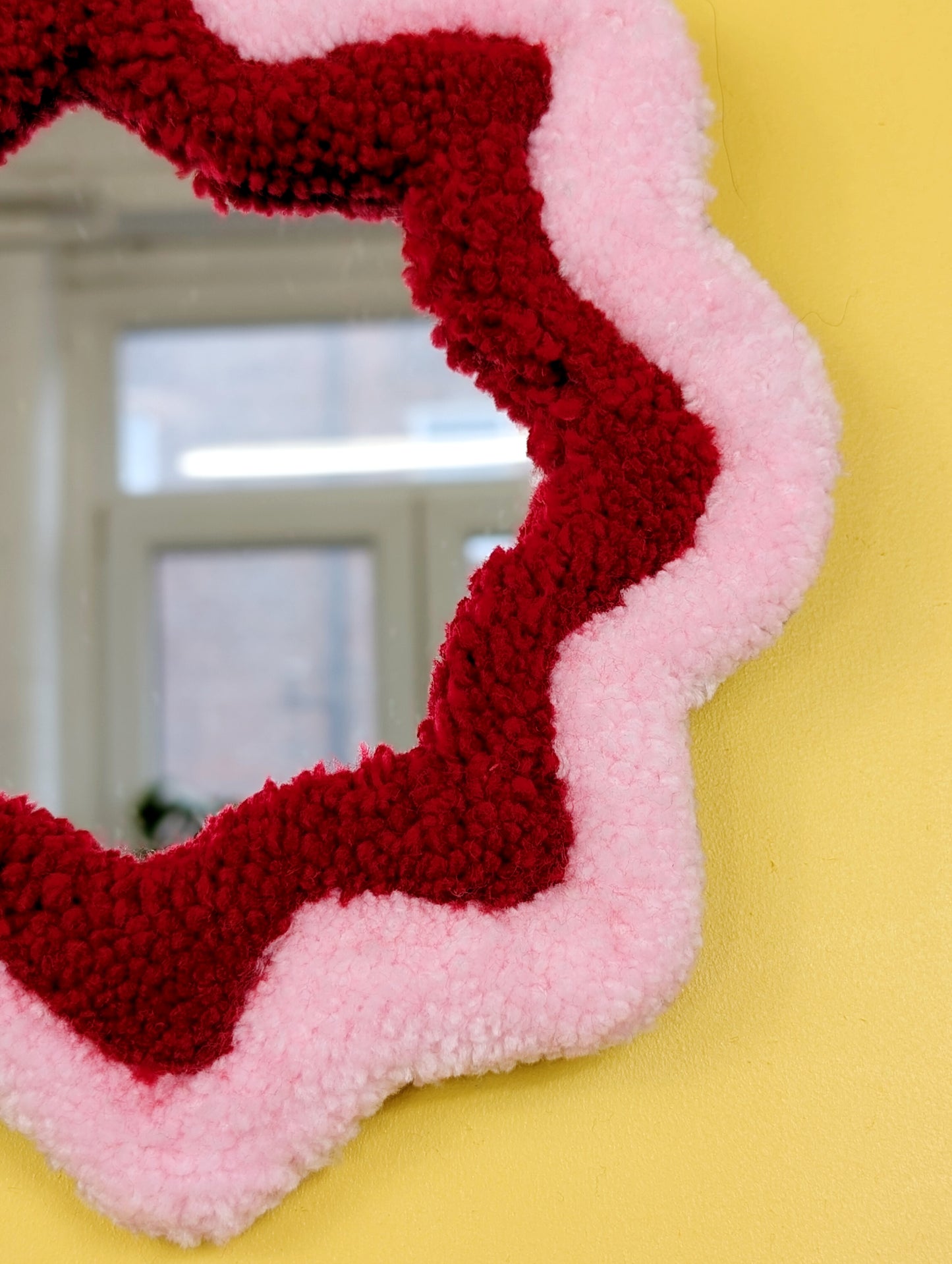  Handmade pink and red tufted mirror with organic wavy shape hanging on a yellow wall.