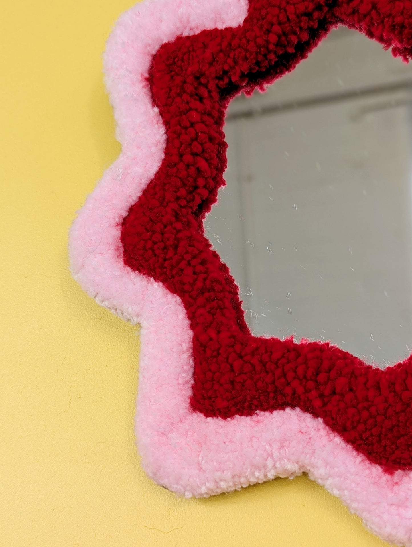  Handmade pink and red tufted mirror with organic wavy shape hanging on a yellow wall.