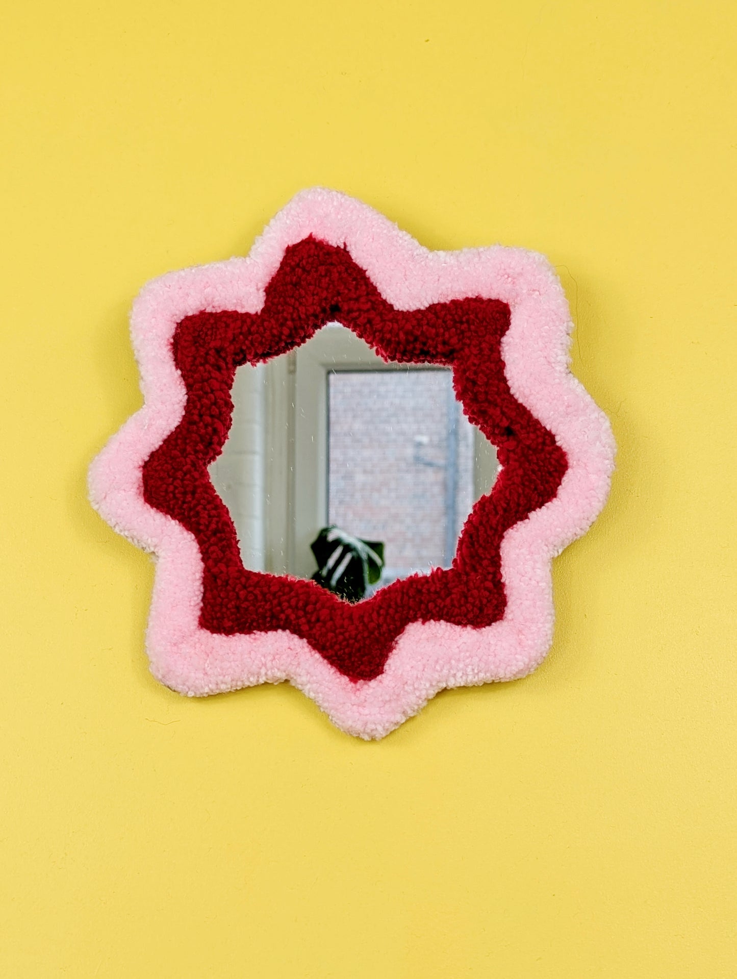  Handmade pink and red tufted mirror with organic wavy shape hanging on a yellow wall.