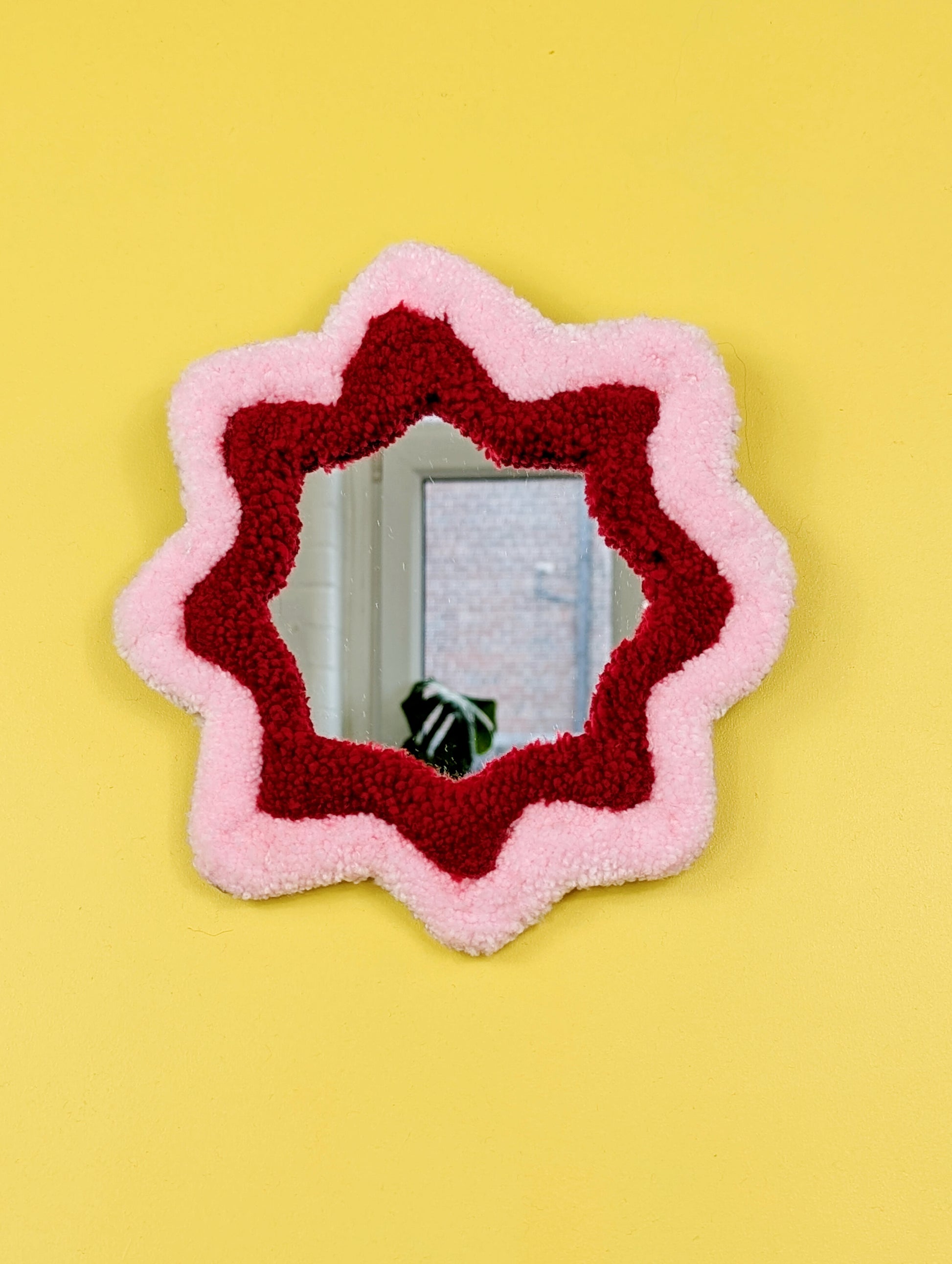  Handmade pink and red tufted mirror with organic wavy shape hanging on a yellow wall.