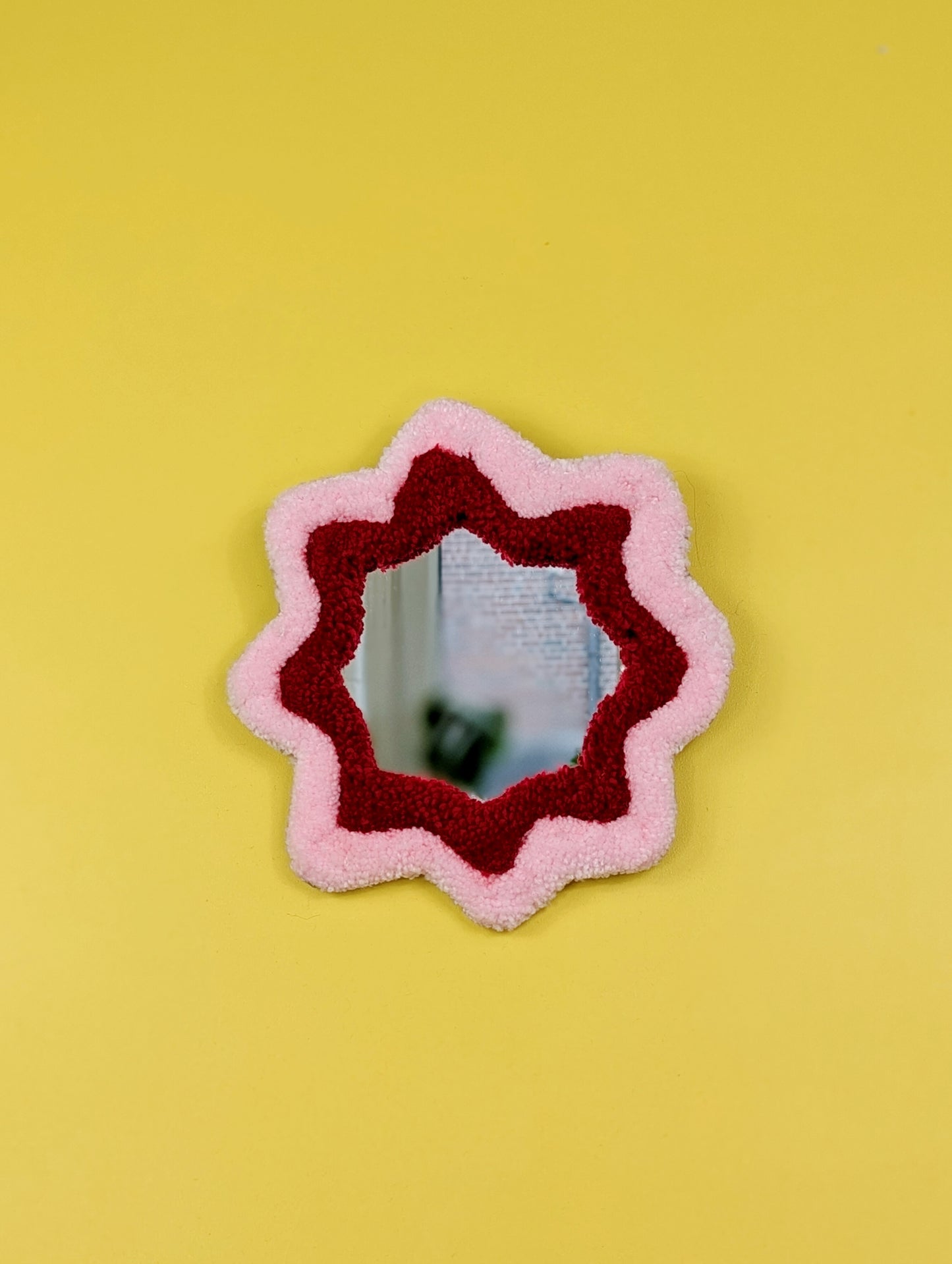  Handmade pink and red tufted mirror with organic wavy shape hanging on a yellow wall.