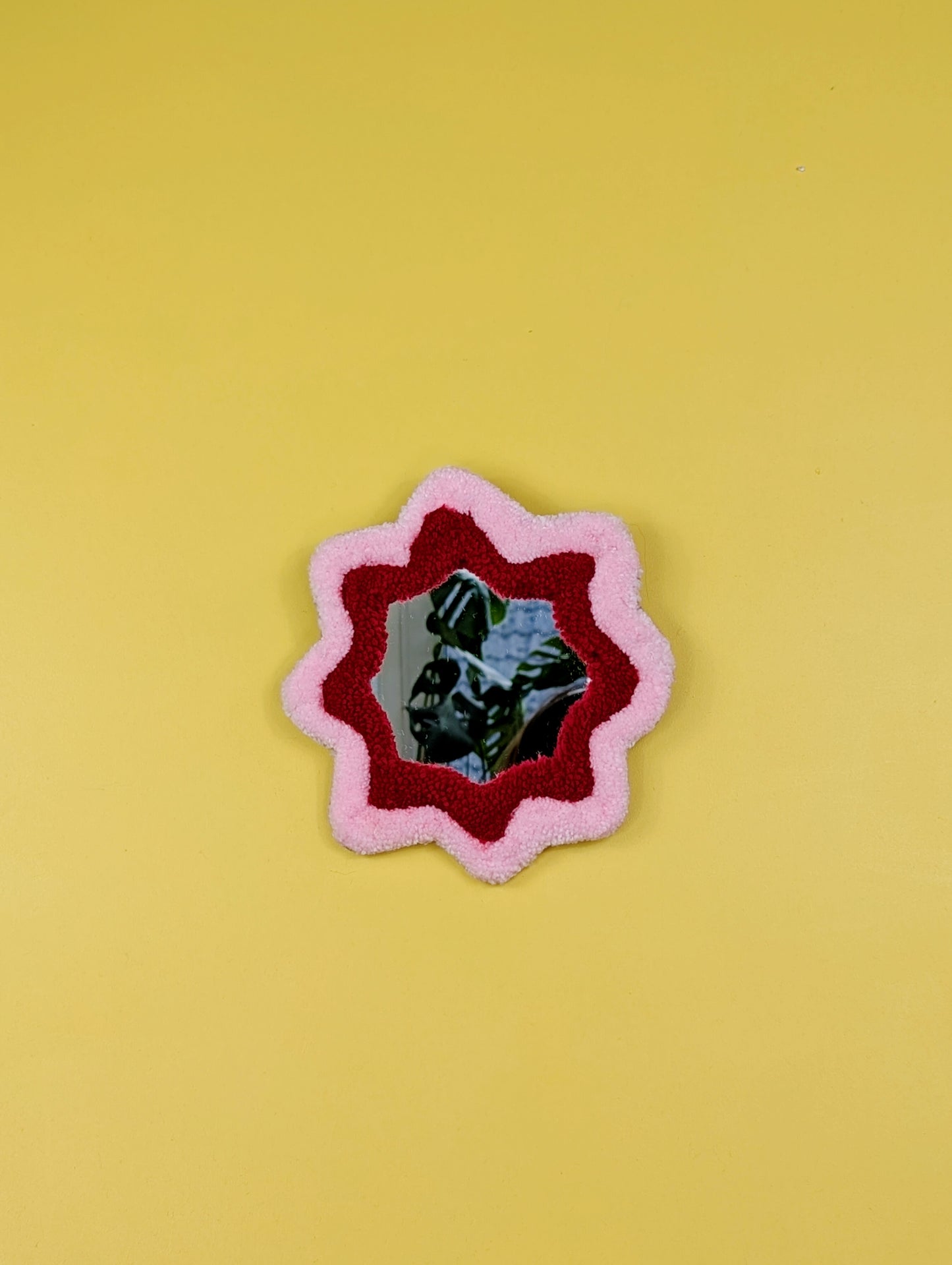  Handmade pink and red tufted mirror with organic wavy shape hanging on a yellow wall.