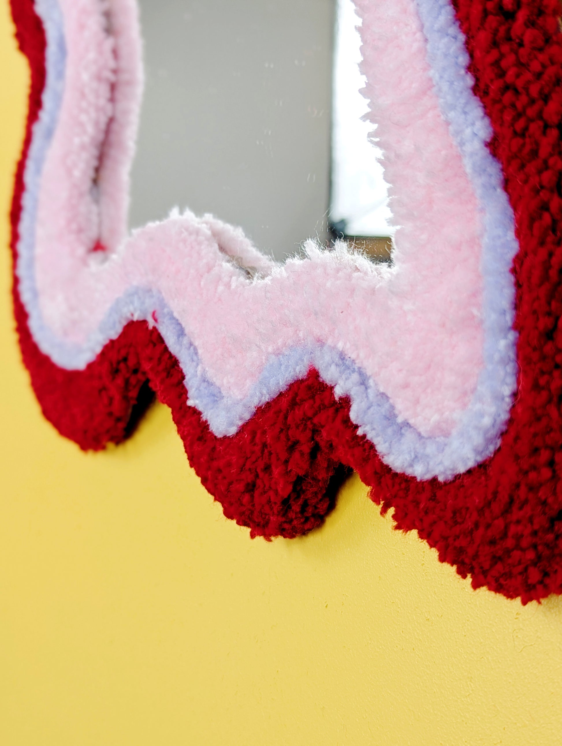 Handmade wavy tufted mirror with colourful textured frame in alternating shades of bright red, pale purple and pale pink, modern aesthetic wall décor for home interiors.