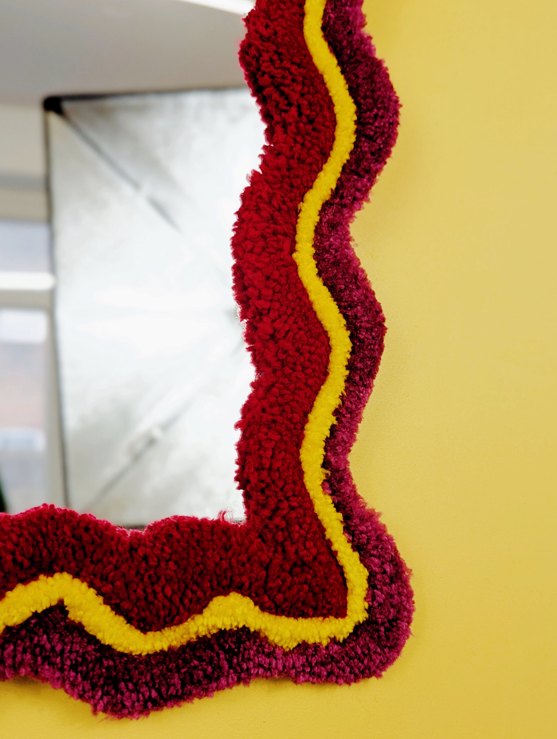 Handmade wavy tufted mirror with colourful textured frame in dark pink, bright yellow and red, modern aesthetic wall décor for home interiors.