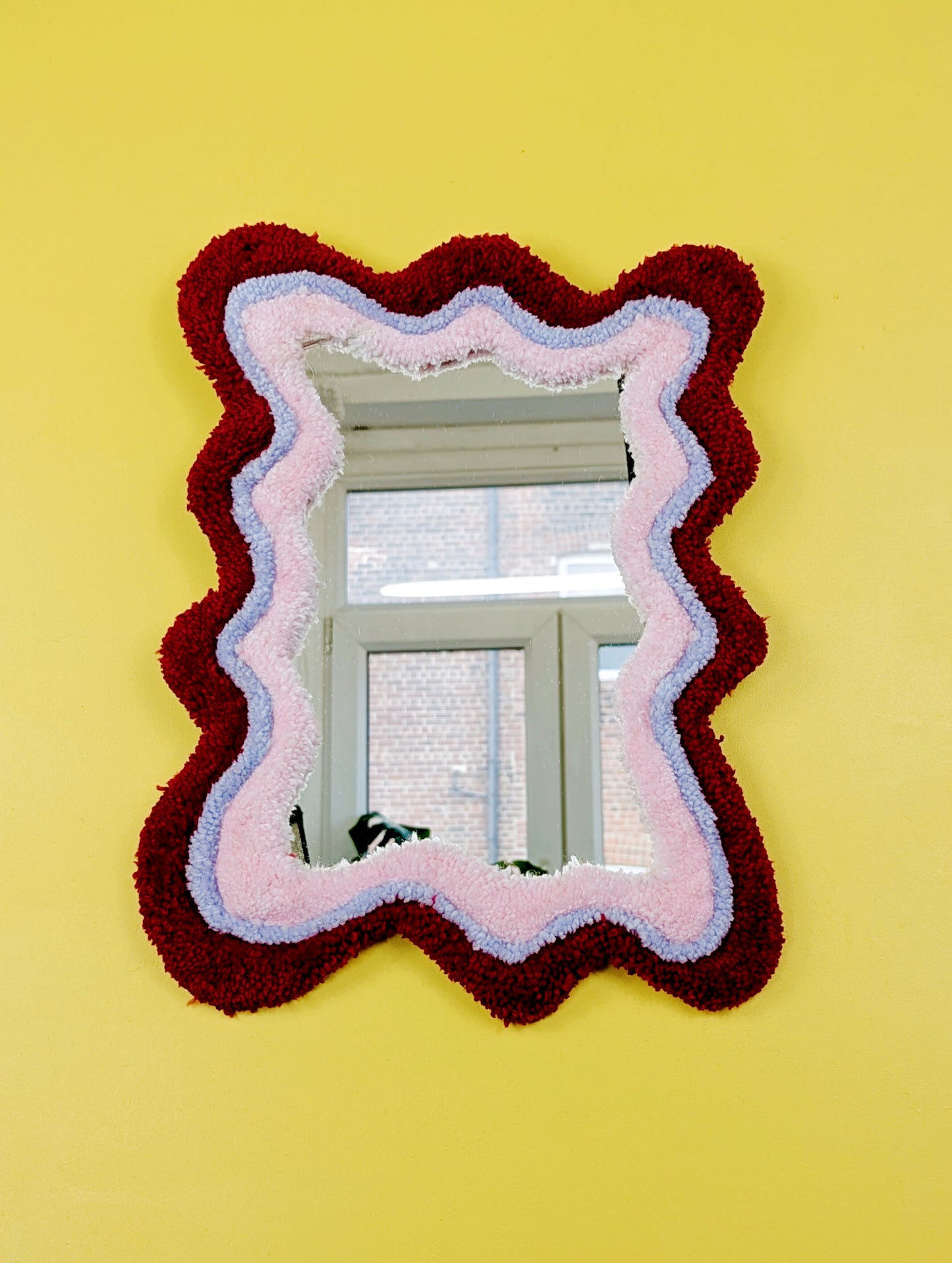 Handmade wavy tufted mirror with colourful textured frame in alternating shades of bright red, pale purple and pale pink, modern aesthetic wall décor for home interiors.