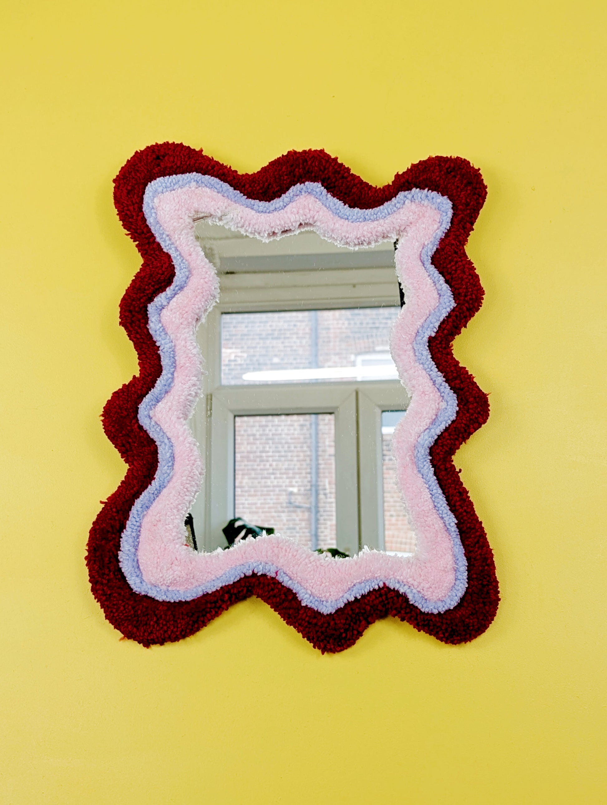 Handmade wavy tufted mirror with colourful textured frame in alternating shades of bright red, pale purple and pale pink, modern aesthetic wall décor for home interiors.