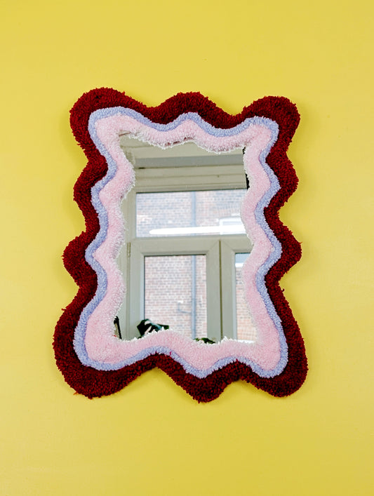 Handmade wavy tufted mirror with colourful textured frame in alternating shades of bright red, pale purple and pale pink, modern aesthetic wall décor for home interiors.