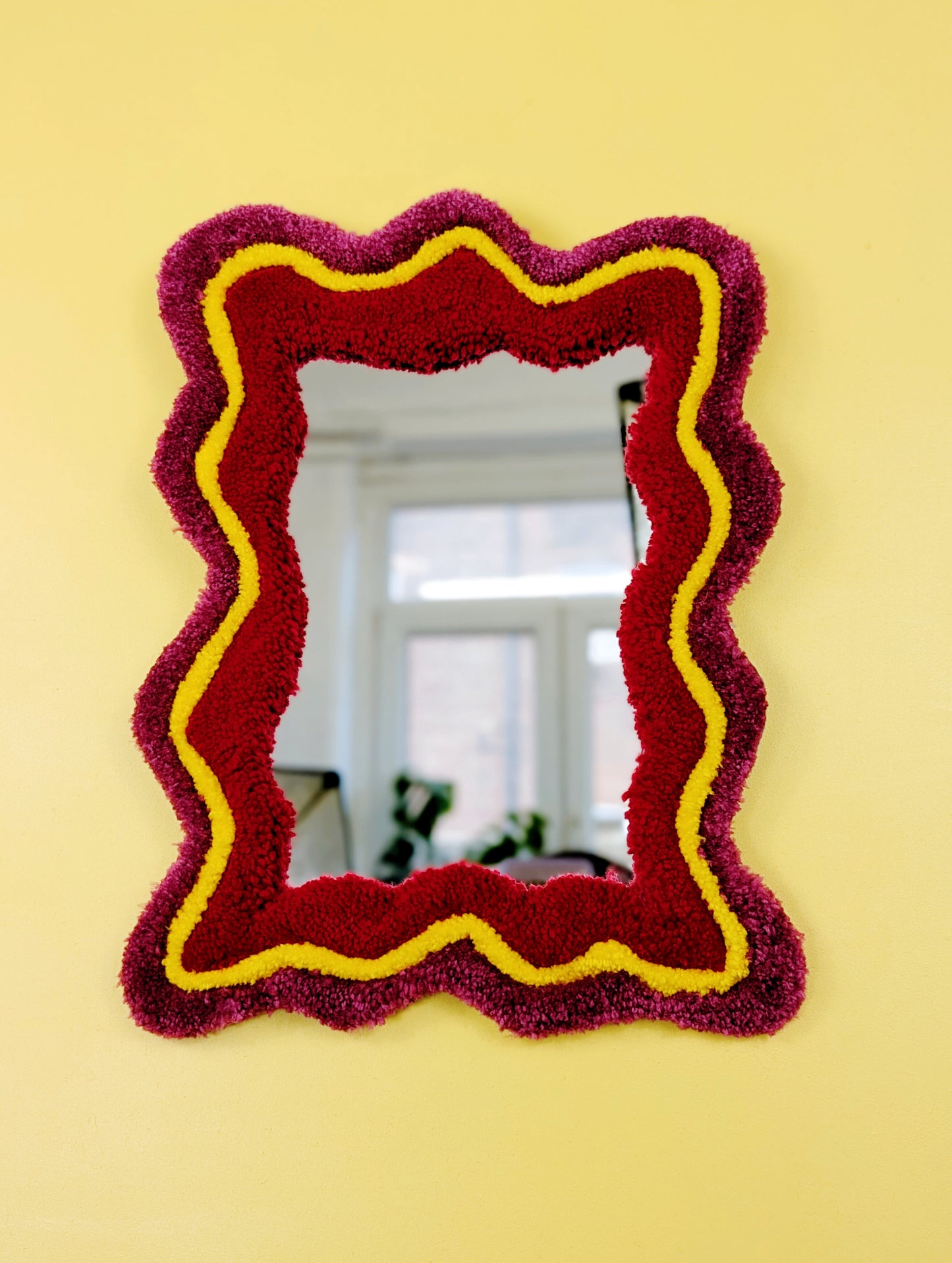 Handmade wavy tufted mirror with colourful textured frame in dark pink, bright yellow and red, modern aesthetic wall décor for home interiors.