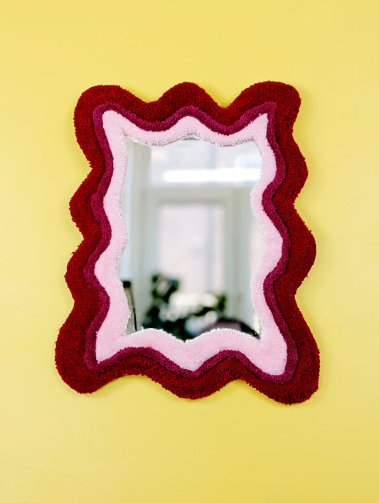 Handmade wavy tufted mirror with colourful textured frame in alternating shades of bright red, dark pink and pale pink, modern aesthetic wall décor for home interiors.