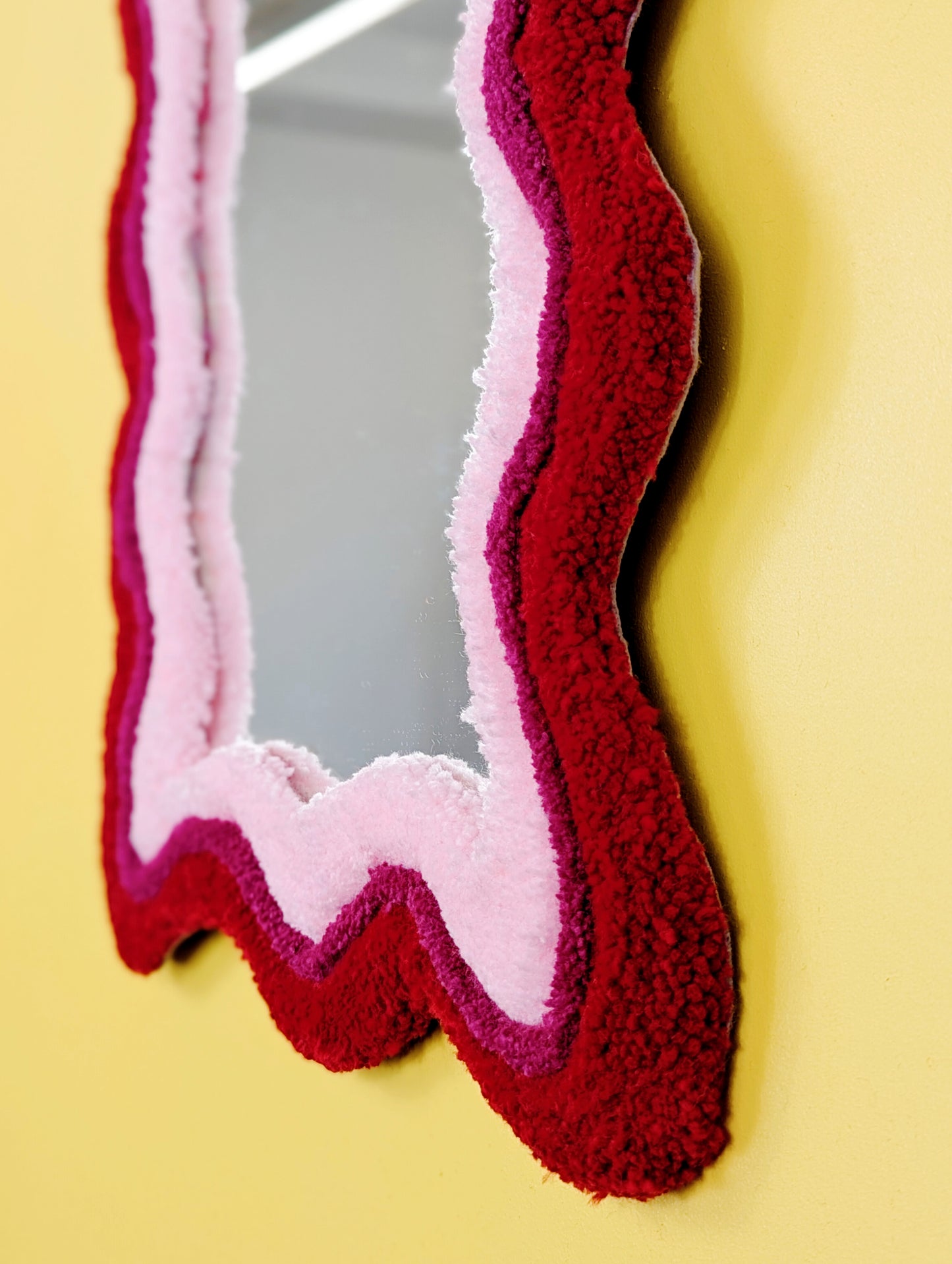 Handmade wavy tufted mirror with colourful textured frame in alternating shades of bright red, dark pink and pale pink, modern aesthetic wall décor for home interiors.
